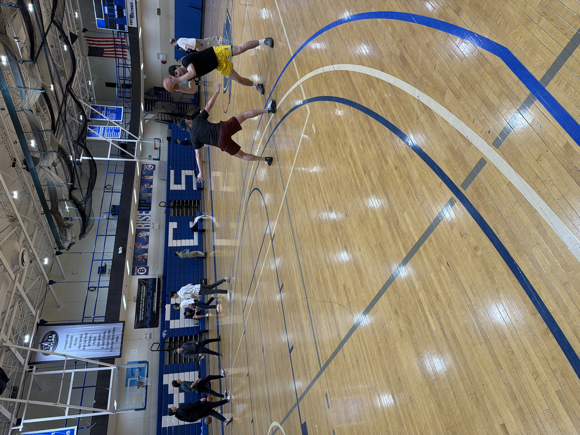 The image shows the interior of a gymnasium, most likely a high school or college basketball court, in what appears to be New York, USA. Two men are practicing a basketball drill on the wooden court, which is marked with blue and white lines. Along the sidelines, multiple people are lined up, holding basketballs. Banners and scoreboards are visible, indicating a school or sports facility. There is an American flag visible. The overall environment suggests a practice session or training activity.