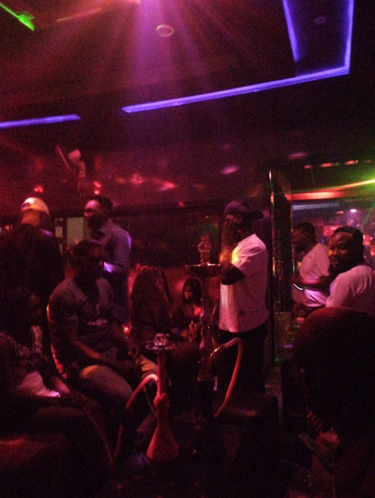 The image shows a dimly lit indoor space in what appears to be a nightclub or lounge. The lighting scheme employs red, purple, green, and blue neon and spot lighting. Several people are present, seemingly engaged in conversation or relaxation. Some individuals are standing, while others are seated. Hookahs are present, suggesting that the setting is one where hookah smoking is permitted. The image provides no direct evidence linking the scene to Gombe, Nigeria. Additional context would be needed to definitively associate the image with a specific location.