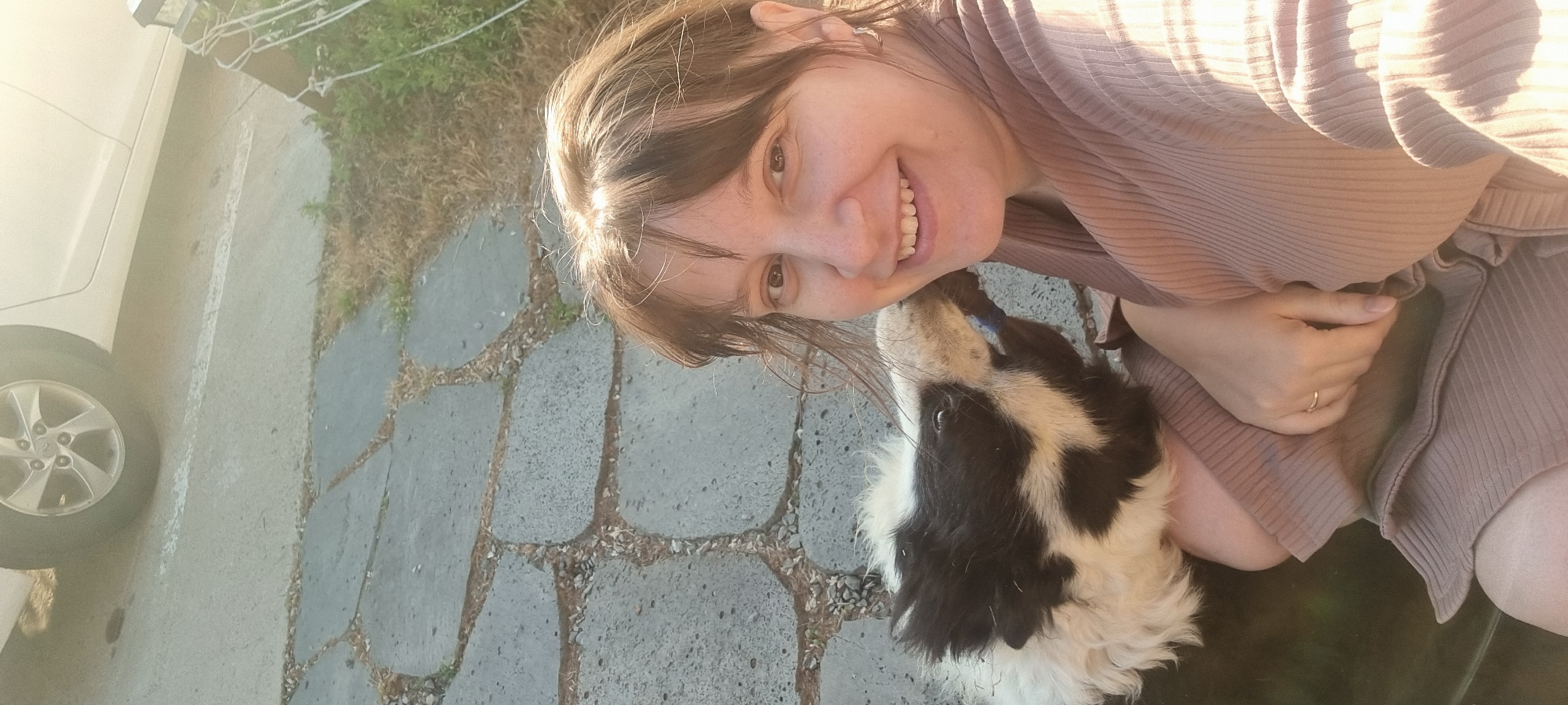 A woman is taking a selfie with a black and white dog on a driveway paved with gray stones, next to a white car. The location is Dunnae-myeon, South Korea. The woman has fair skin and is wearing a beige ribbed shirt or dress. She is smiling at the camera. The dog is looking up towards the woman.