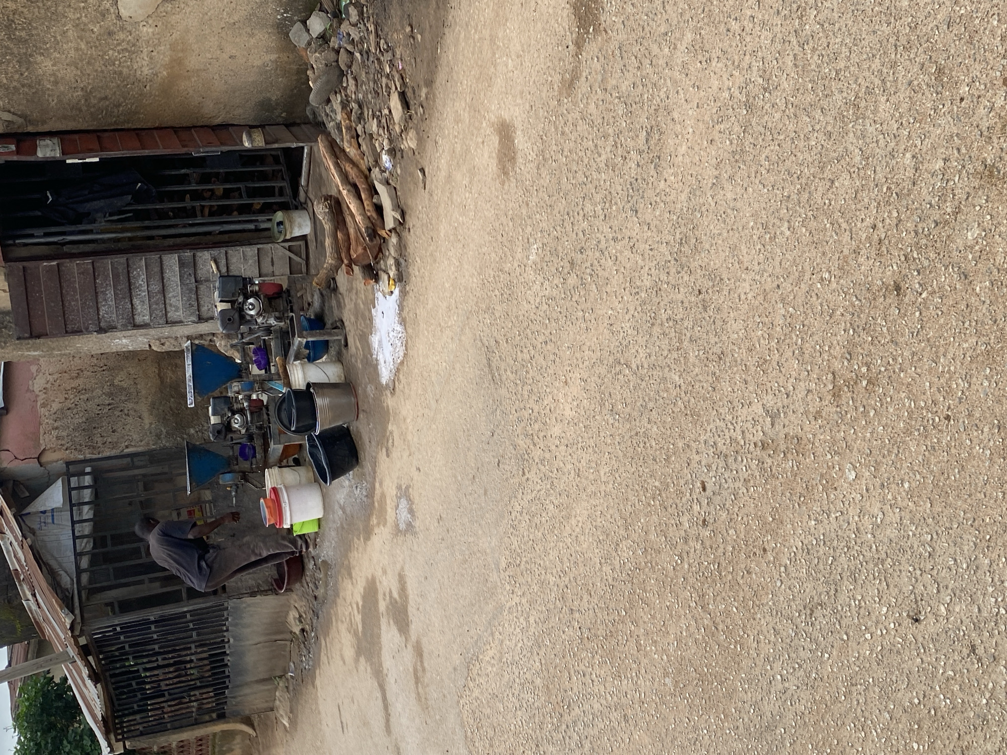 In the city of Jos, Nigeria, an outdoor scene depicts a person dressed in dark clothing tending to two agricultural processing machines positioned near a doorway. The machines, likely for grinding or processing crops, are blue and silver, with visible funnels and engines. Several buckets, some containing liquids, are scattered around the machines and on the ground. In the foreground, a rough, gravelly surface of a road or pathway occupies the majority of the frame. To the right, a pile of cut logs and stones are visible. The background reveals a weathered building with a corrugated metal roof and a wooden door. The overall environment suggests an informal, potentially commercial, setting for agricultural activities.