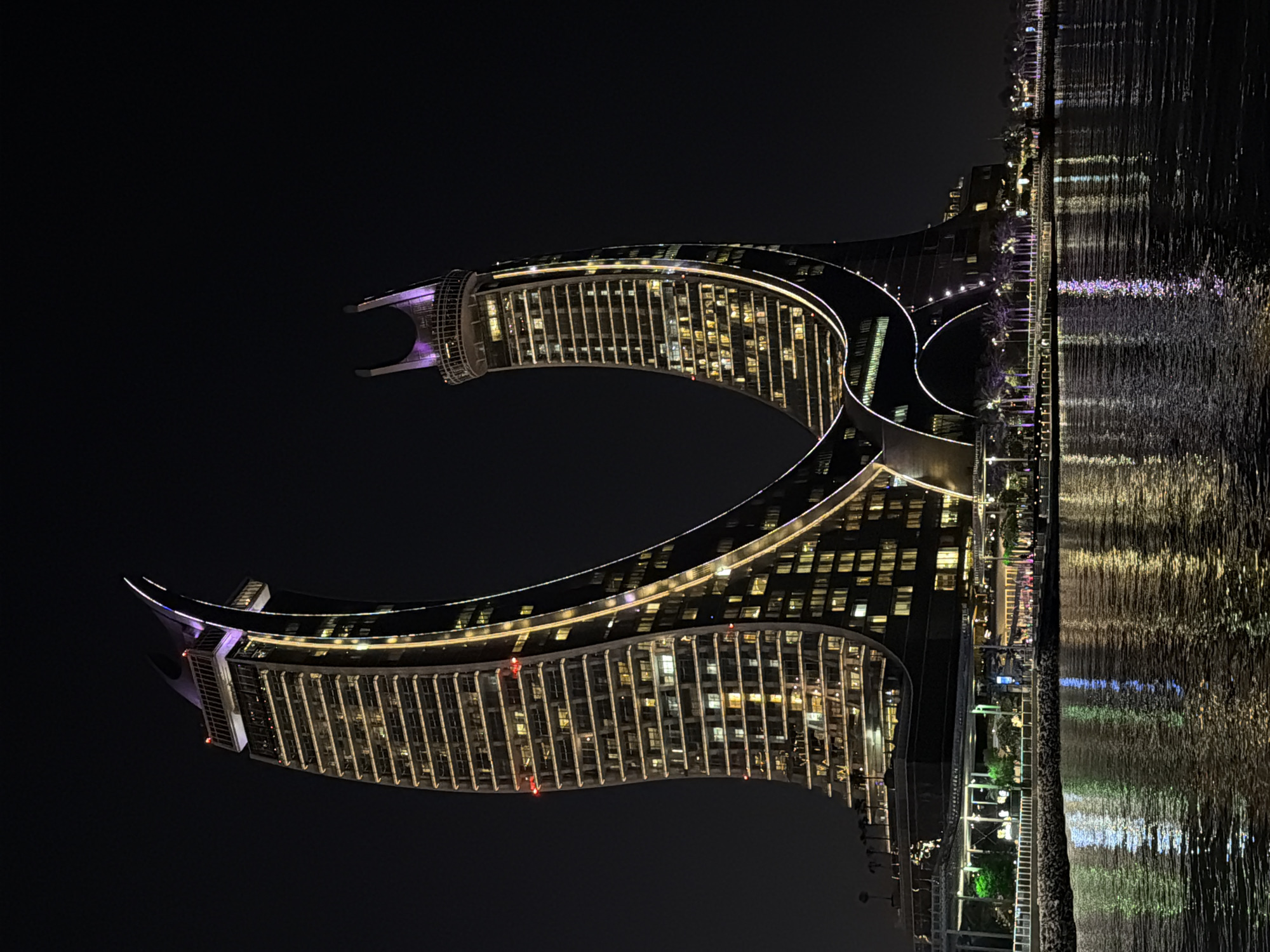 An exterior, eye-level, full shot at night shows two curved, modern skyscrapers with illuminated windows and lights along their edges. The buildings are dark brown and gold, with some purple lighting at their peaks. The skyscrapers are positioned close together, with their curved structures nearly touching in the middle, creating a V-shape. The buildings reflect in the dark water in front of them, creating streaks of gold, white, and purple light on the surface. A promenade with palm trees and some green lighting runs along the base of the buildings. The sky is completely dark.