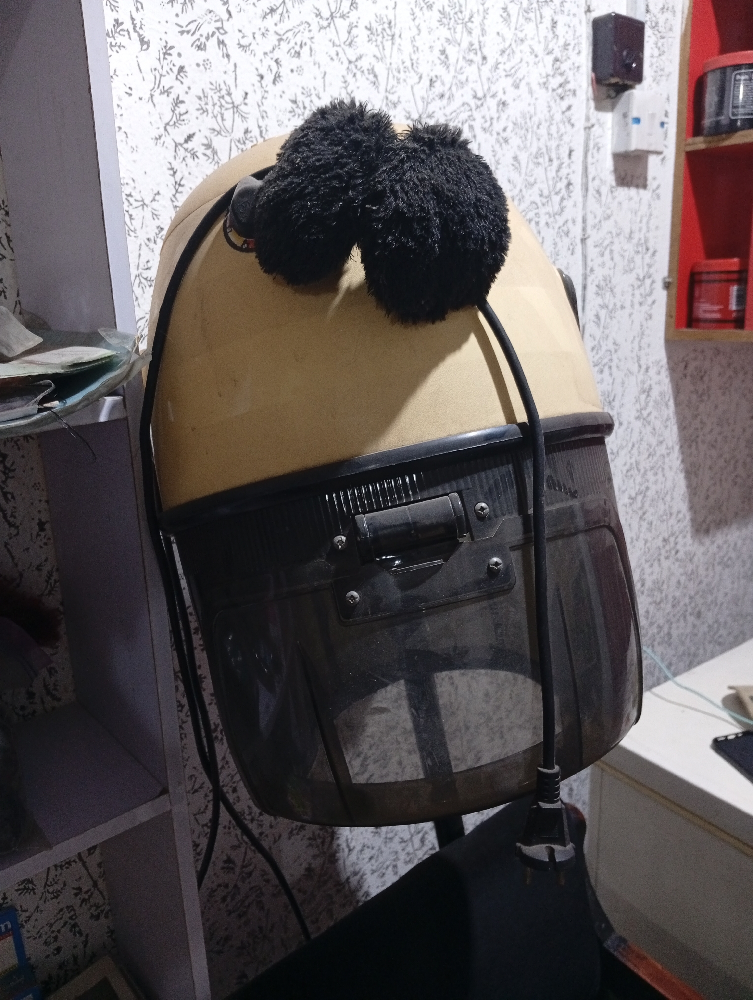 A hair dryer sits on a chair in a room with patterned wallpaper. The hair dryer is beige with a black base, and a pair of fluffy black earmuffs are positioned on top of it. A black electrical cord descends from the hair dryer, ending in a three-prong plug. To the left is a white shelving unit, and to the right is a white cabinet. A red shelf unit with various objects is visible on the wall. Based on the power plug configuration, and the prompt information, the location is Lokoja, Nigeria.