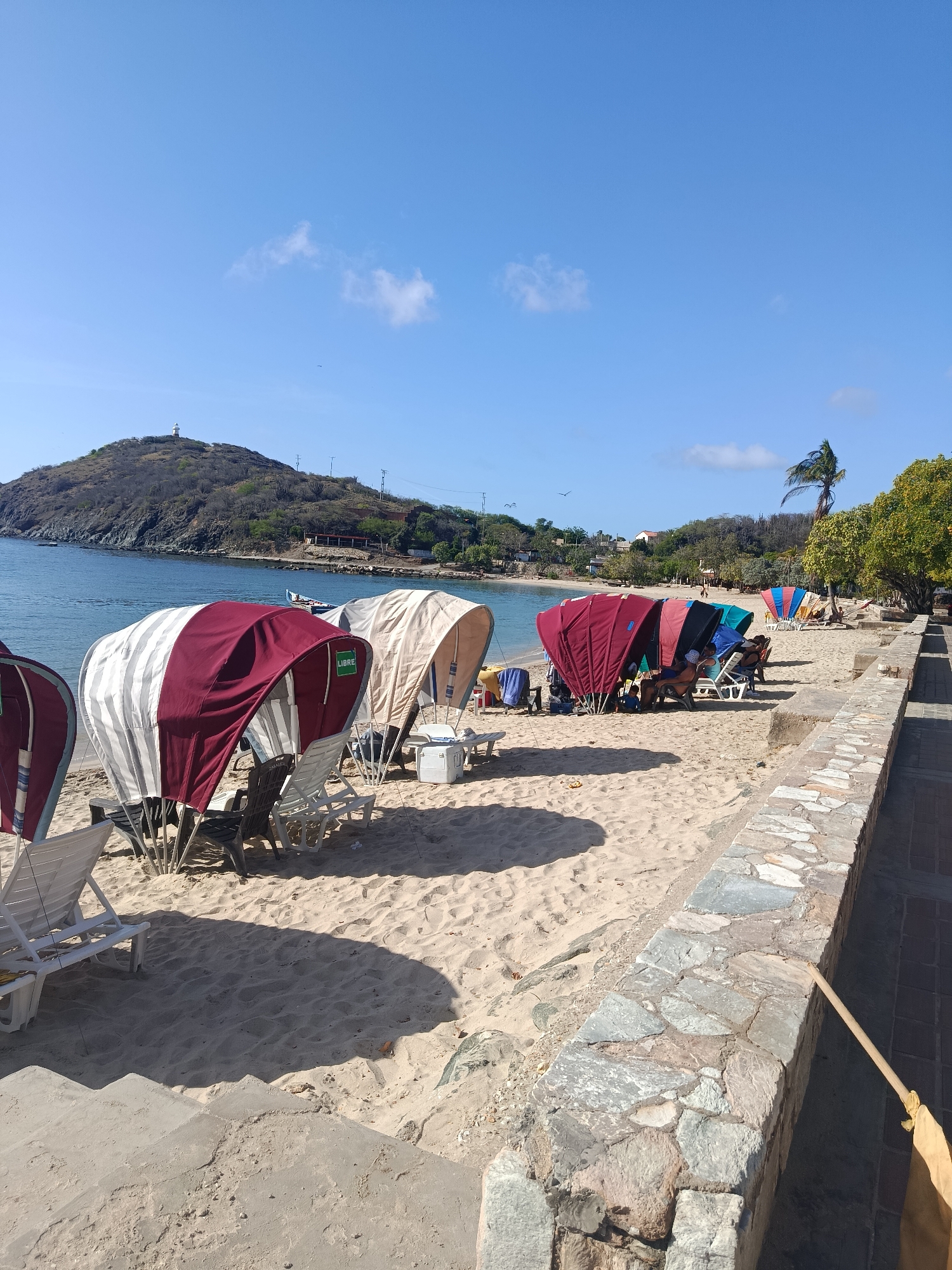 In Pedro González, Venezuela, a sunny beach scene unfolds with a clear blue sky and a calm body of water. Foreground features a sandy expanse with several beach tents providing shade. One tent is red and white striped, with a white cooler next to it and several white lounge chairs partially visible. Further down the beach, a row of tents in various colors, including red, blue, and green, are set up, with people seated in lounge chairs underneath them. A stone wall borders the beach, with a paved walkway behind it. In the background, a lush green hill rises from the water, topped with a white lighthouse. Trees and some buildings are scattered along the coastline. The overall atmosphere is one of leisure and relaxation on a tropical day.