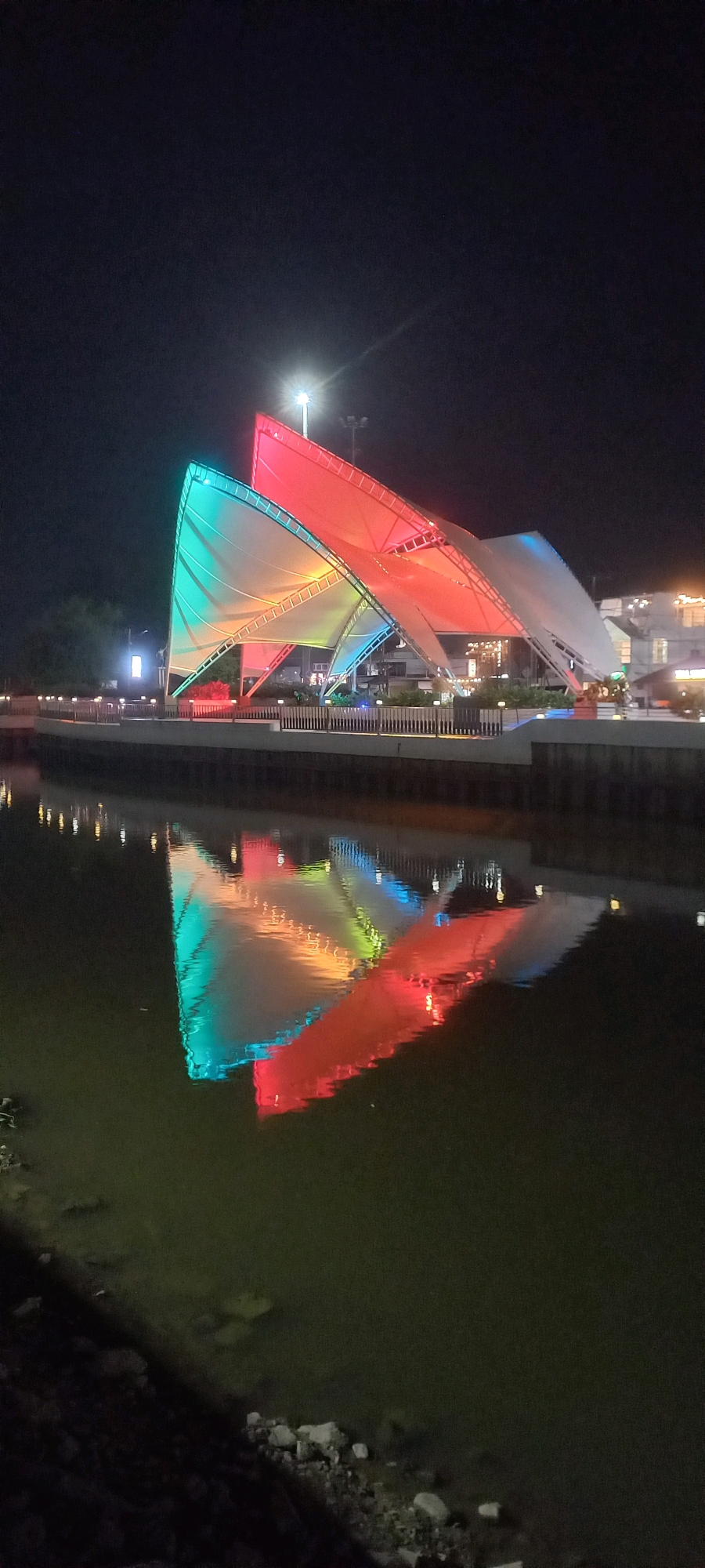 A nighttime, vertical, medium shot captures a modern, sail-like structure illuminated with vibrant, changing lights against a dark sky. The structure, featuring large, angled white fabric panels, is colored with red, green, and blue lights. A bright spotlight is visible at the top of the structure. A railing and walkway border the structure, separating it from a dark body of water in the foreground. The water reflects the colorful lights of the structure. The scene appears to be a public space or waterfront in Melayu, Indonesia.