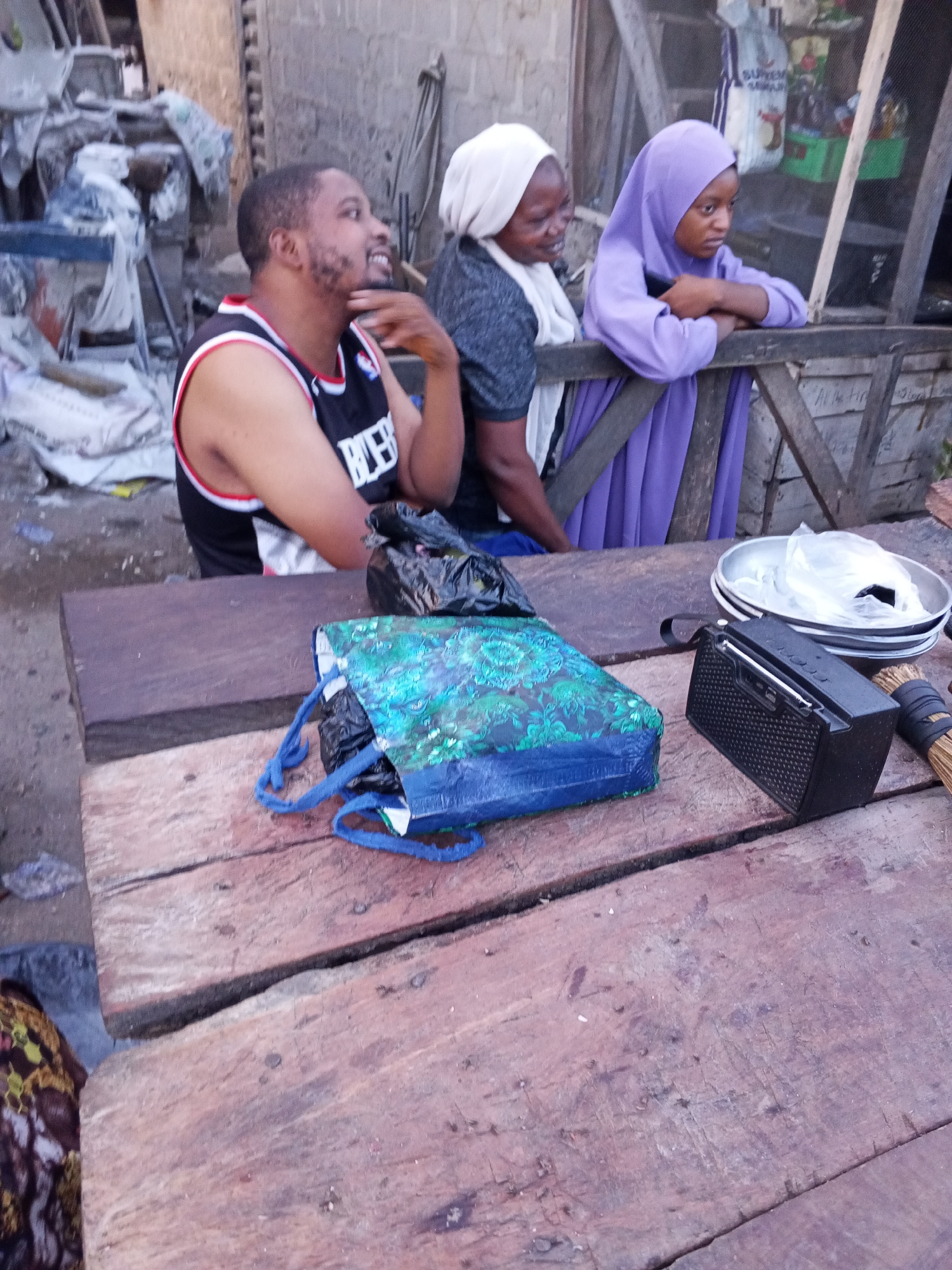 The scene is set in Zango-ndaji, Nigeria. Three individuals are present: a man wearing a basketball jersey seated at a wooden table, and two women, one in a white head covering and the other in a lavender hijab, leaning against a wooden structure. On the table, there is a blue patterned shopping bag, a black plastic bag, a small black radio, a stack of metal bowls, and a small bundled broom. The background shows a cluttered outdoor area with various items, including bags and building materials. The table appears to be constructed from weathered planks.