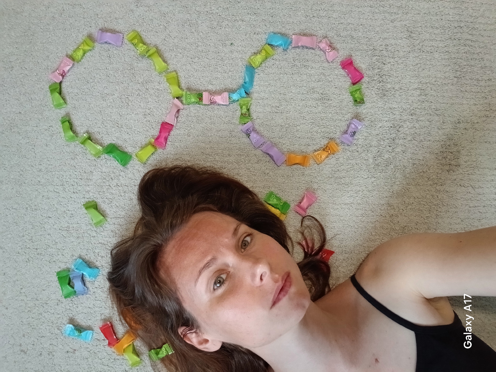 A woman with brown hair lies on a light-colored carpet. Arranged around her head are colorful, individually wrapped candies, forming a pair of glasses shape. The scene is lit by natural light. A black spaghetti strap is visible on her shoulder. A watermark that says "Galaxy A17" is printed at the lower right of the photo. This image was not taken in Yekaterinburg, Russia.