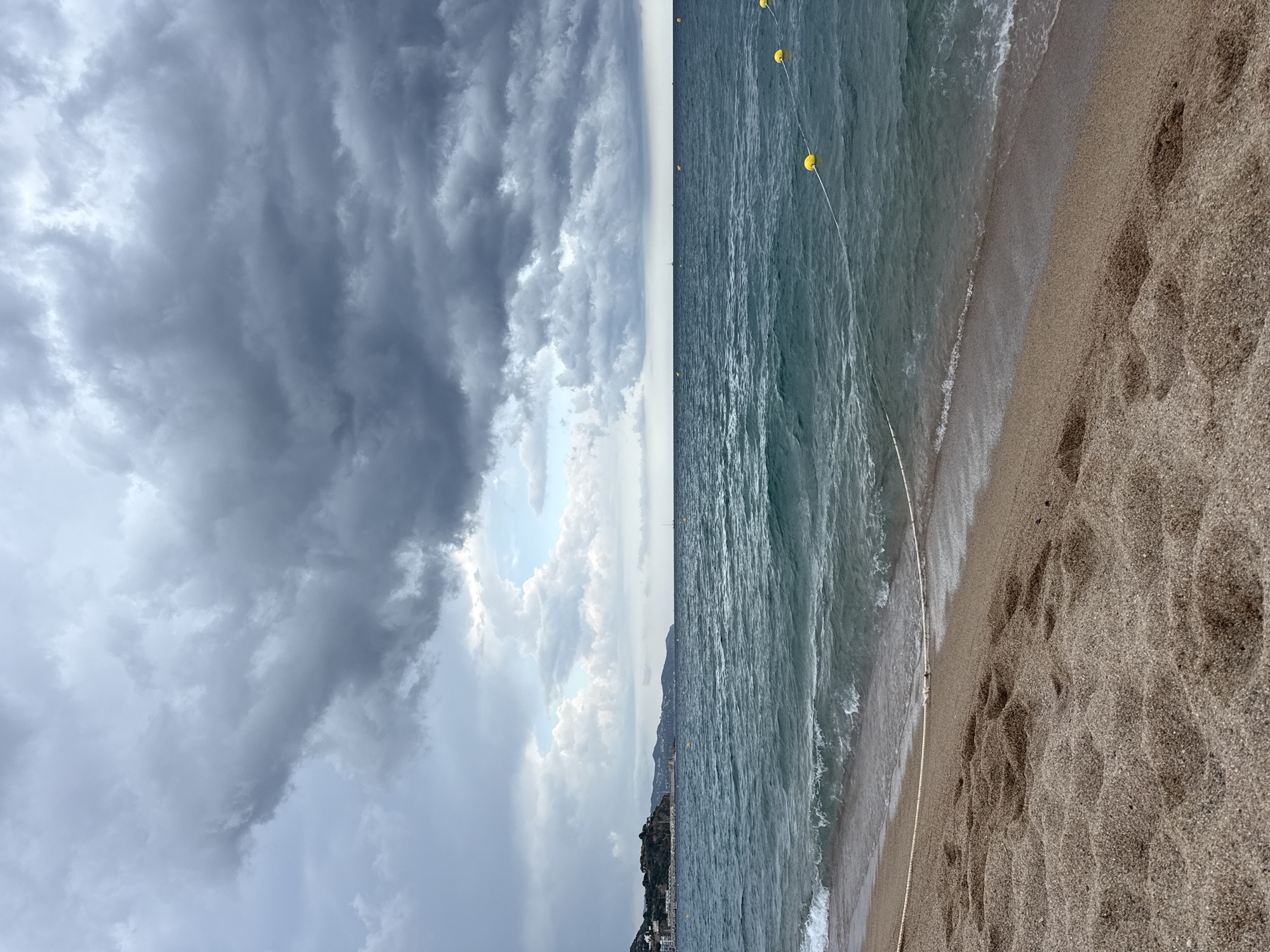 The image depicts a beach scene in Blanes, Spain, with a cloudy sky overhead. The foreground is dominated by sandy beach with footprints visible. The midground features the turquoise sea with gentle waves lapping the shore, and a white rope with yellow buoys marking a section of the water. In the background, the coastline is visible with buildings and lush green hills. The overall atmosphere is overcast, suggesting potential rain. There are no people visible in the image.