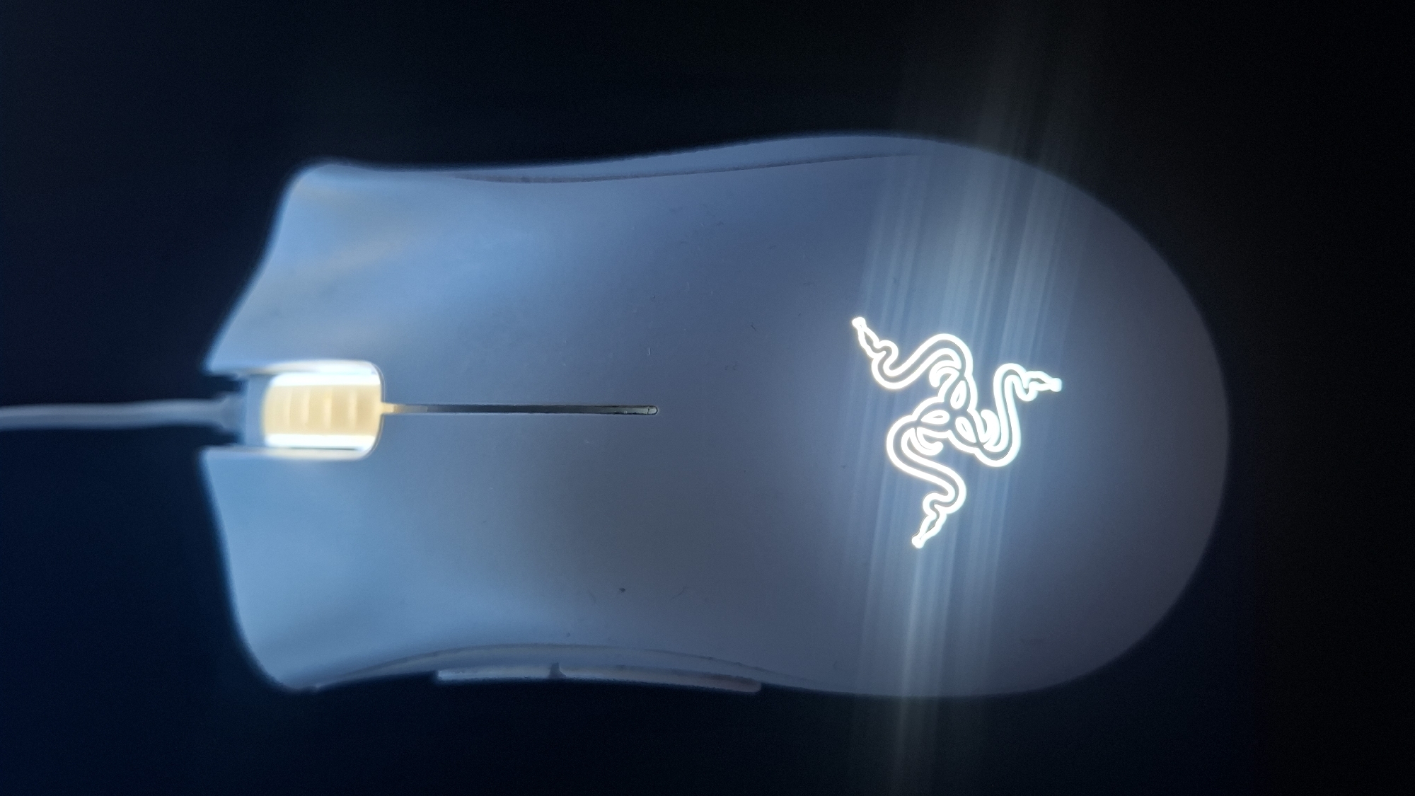 A white Razer gaming mouse is photographed against a dark background. The scroll wheel is illuminated with a yellow-orange light. The Razer logo is lit up brightly on the right side of the mouse. The mouse's cable is visible on the left side.