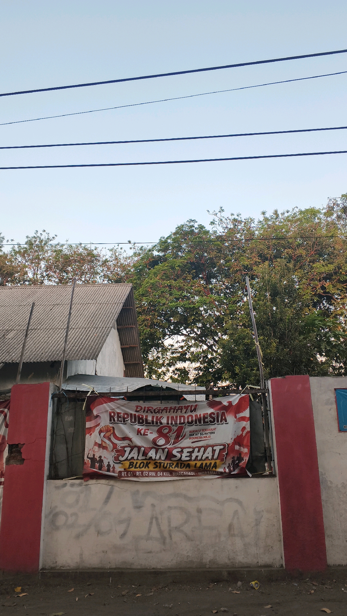A banner celebrating the 81st anniversary of the Republic of Indonesia is displayed on a red and white wall in Indramayu, Indonesia. The banner features the Indonesian flag, a Garuda bird symbol, and the text "DIRGAHAYU REPUBLIK INDONESIA KE-81 JALAN SEHAT BLOK STURADA LAMA". Below the banner, graffiti is visible on the wall. Trees and a building with a corrugated roof are present in the background, and power lines run across the top of the image. The scene appears to be outdoors on a street.