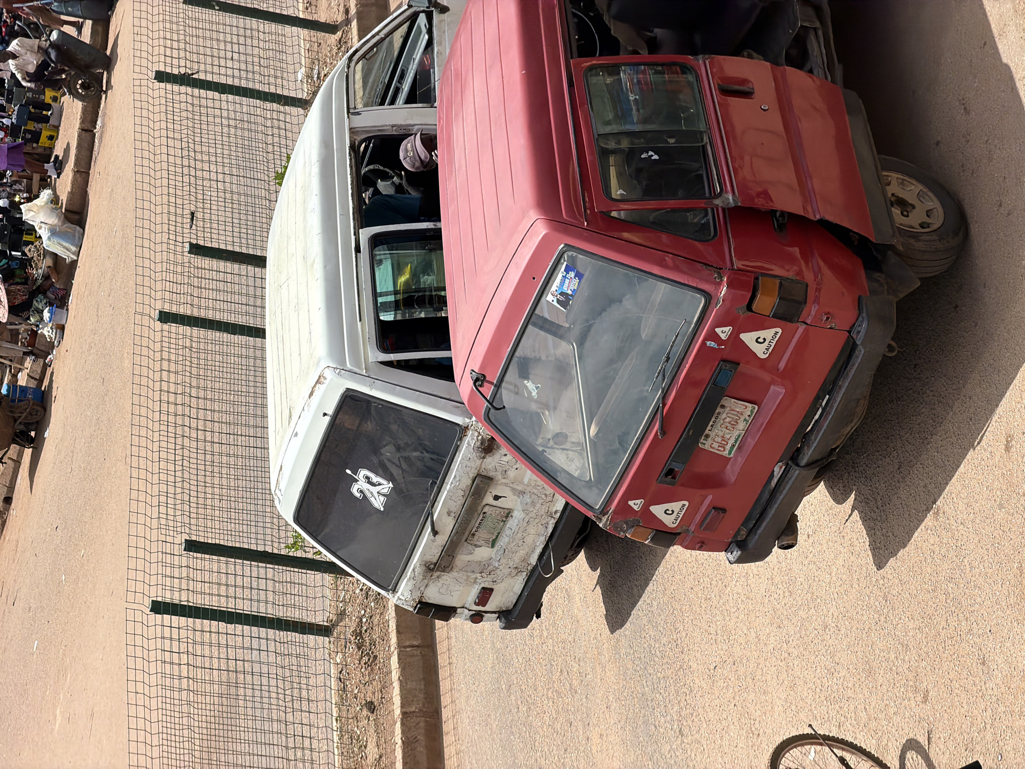 A high-angle, medium shot captures two utility vans parked on a dirt road in Bukuru, Nigeria. The lead vehicle is a red, three-wheeled van with its passenger door ajar, revealing an empty interior. A sticker with a stylized 'C' in a triangle and the word "CAUTION" is visible on the rear quarter panel. Another similar sticker is present on the driver's side rear. The van has a license plate that reads "LAGOS GCE 860XJ". Behind the red van is a white, four-wheeled van, appearing older and weathered, with extensive peeling paint. A sticker of the number "23" with a basketball player silhouette is on its rear window. The van's rear license plate is obscured. The background shows a dirt lot with a wire fence and a busy market scene, including people and various stalls. The overall lighting suggests a bright, daytime setting.