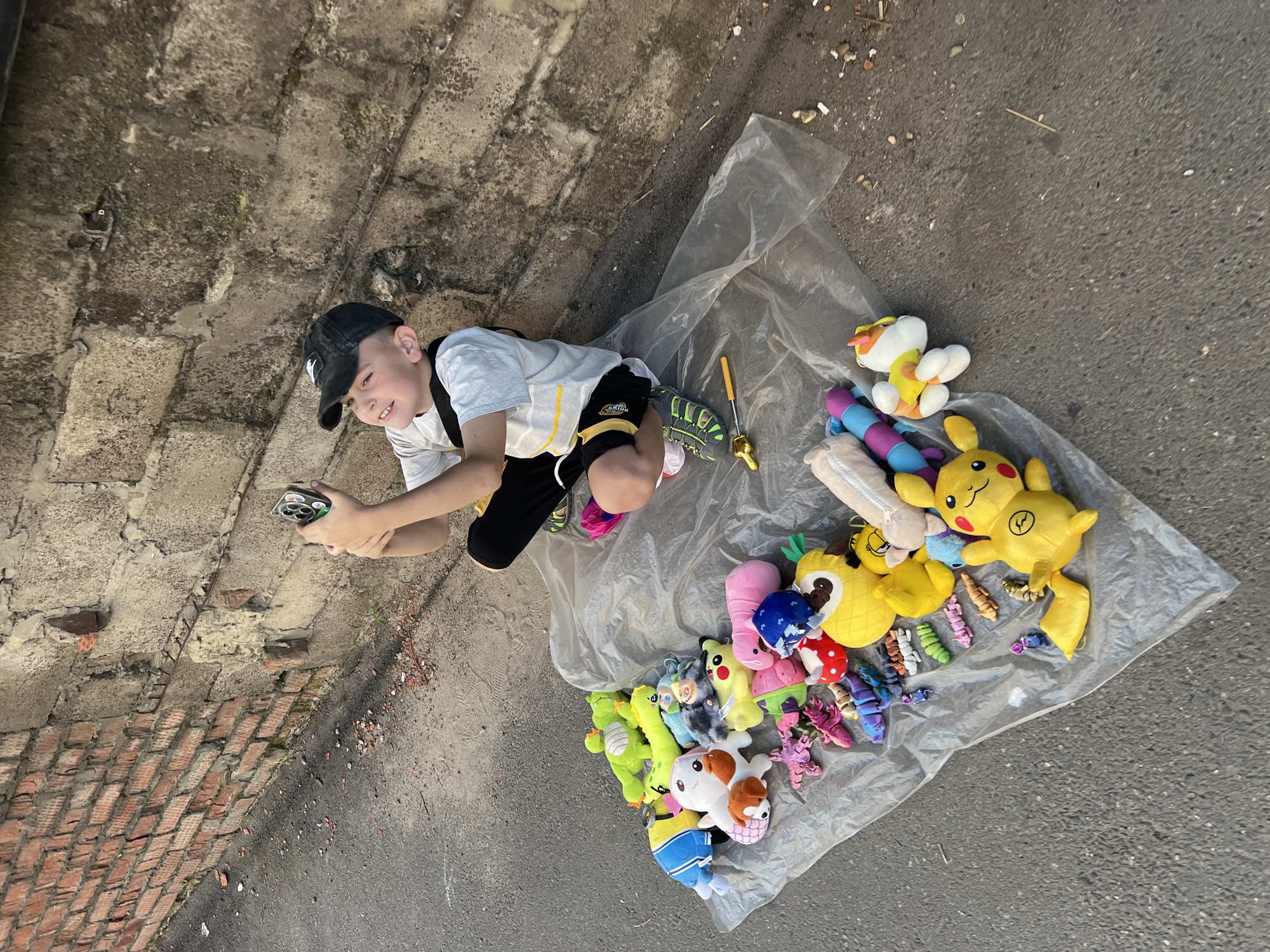 In Krasnodar, Russia, a young boy wearing a black baseball cap and a light-colored t-shirt with black shorts is lying on the ground. He is looking at a smartphone in his hands. Scattered around him on a clear plastic sheet is a collection of colorful stuffed toys, including several Pokémon characters like Pikachu, as well as various other plush animals and what appear to be small articulated toys. The setting is outdoors, with a rough concrete surface and some brickwork visible in the background. The scene suggests a casual, possibly recreational moment, with the boy engaged with his device amidst his collection of toys. The lighting indicates it is daytime, likely sunny, given the overall brightness. There are no visible weather cues, but the ground appears dry.