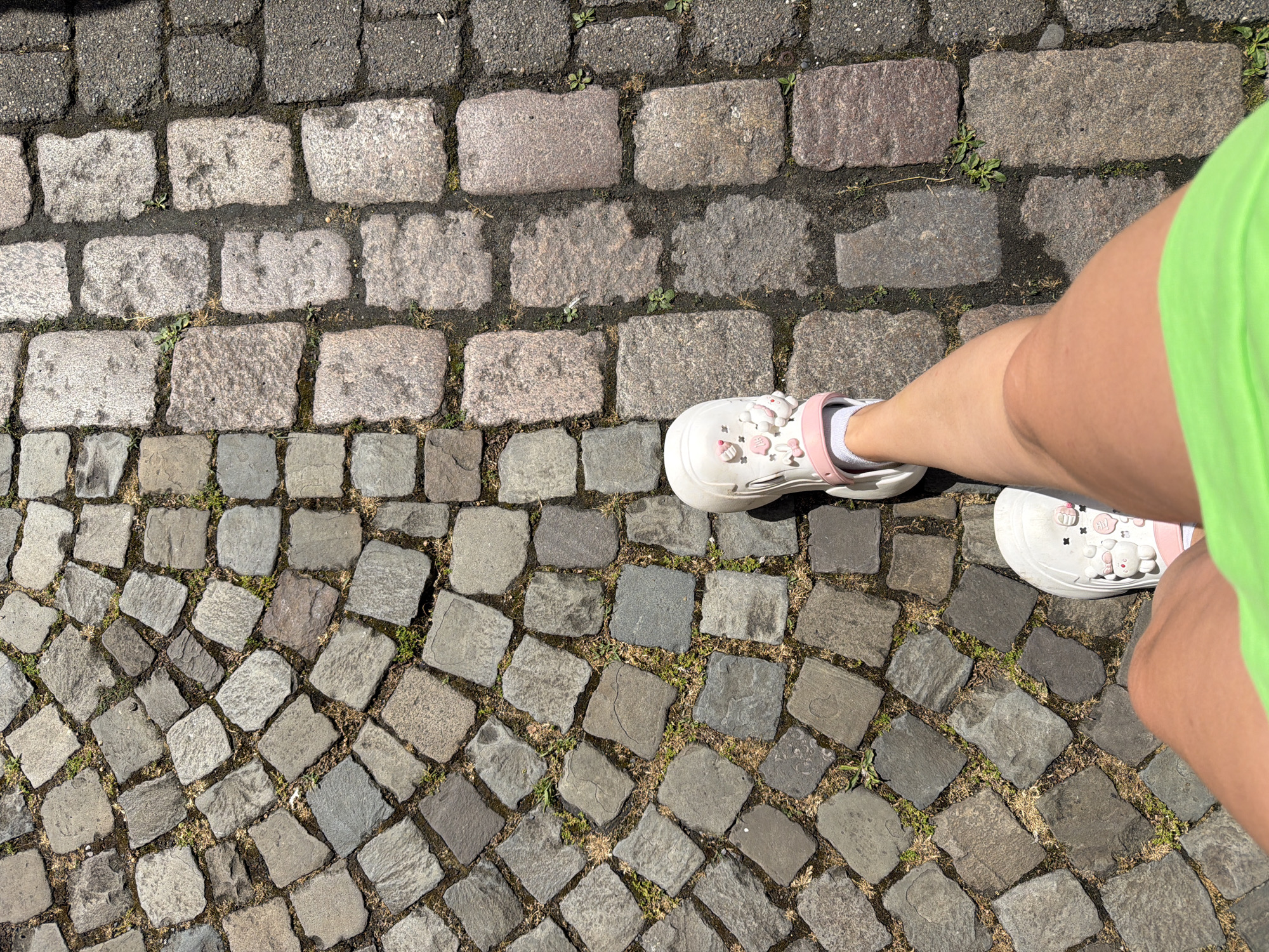 An overhead, eye-level shot captures a person's legs and feet from the knee down, walking on a cobblestone street. The person is wearing white Crocs with pink accents, white socks, and neon green shorts. The cobblestones transition from small, irregular grey stones to larger, rectangular pink and grey stones, and finally to dark grey asphalt pavers. Green weeds are growing between the stones. The scene is brightly lit by sunlight, casting shadows. The location is identified as Telgte, Germany.