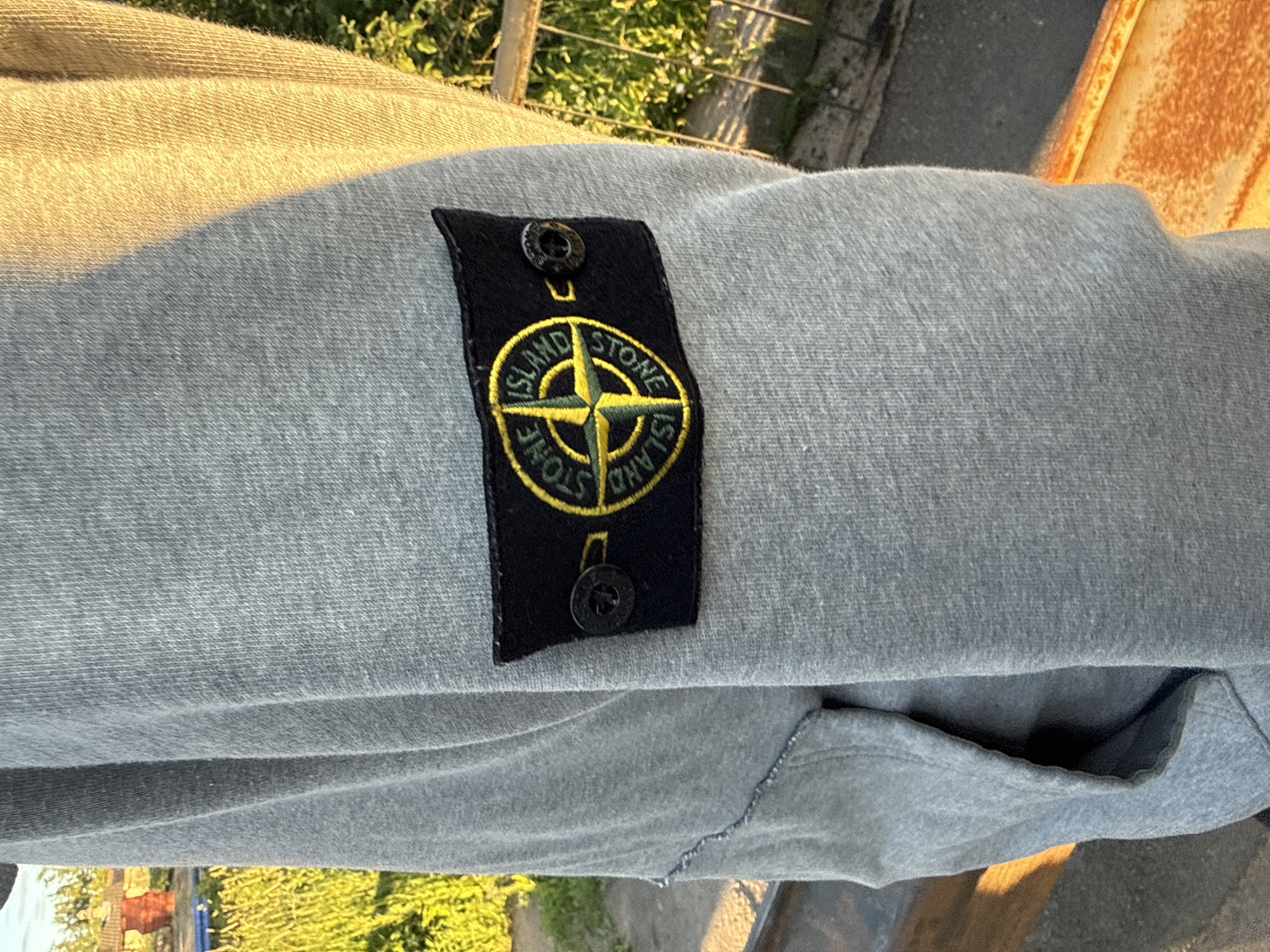 The image shows a close-up of the sleeve of a grey sweatshirt, featuring a black patch with the Stone Island logo. The logo is a compass rose in yellow and green, with "STONE ISLAND" written in a circular pattern. Two black buttons are attached to the patch. The sweatshirt appears to be made of a soft, textured fabric. The background is blurred, but suggests an outdoor setting with greenery and possibly a building. There is no indication of this image being taken in Saint Petersburg, Russia.