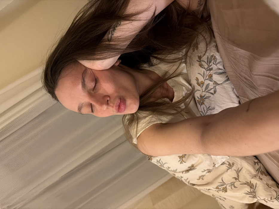 An adult female is depicted in an indoor residential or accommodation setting in Budva, Montenegro. She has long, dark brown hair and wears a light-colored, short-sleeved top. Her eyes are closed, and her lips are puckered. Her head rests on her right hand, which is positioned on a pillow featuring a pattern of green olive branches with yellow-orange berries on a light background. A dark tattoo is visible on her right inner forearm. She is situated on a bed covered by plain, light brown bedding. In the background, light-colored, vertically folded curtains are visible against a beige wall. The scene is illuminated by warm, artificial light, and the individual is posing for a self-portrait.