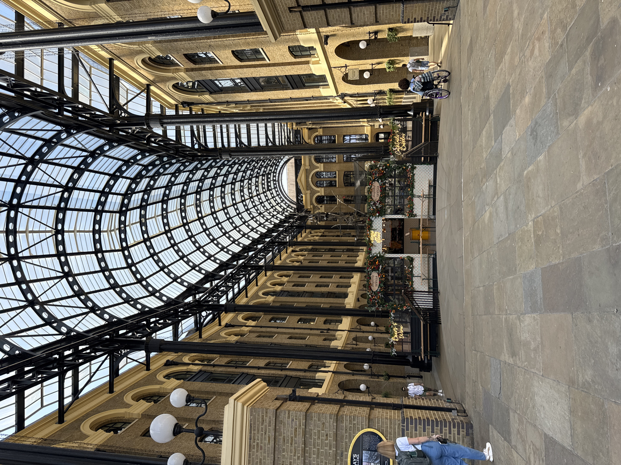 This is an interior view of the Leadenhall Market in the City of London, United Kingdom. The building features a wrought-iron and glass roof, supported by ornate metal beams. The walls consist of multi-story buildings constructed of beige brick with arched windows. There are shops and signage along the walkway, decorated with holiday decorations. People are seen walking, some in casual attire. The ground is paved with rectangular stone tiles. Street lights are attached to the buildings. The image is taken from a low angle, looking towards the center of the market.