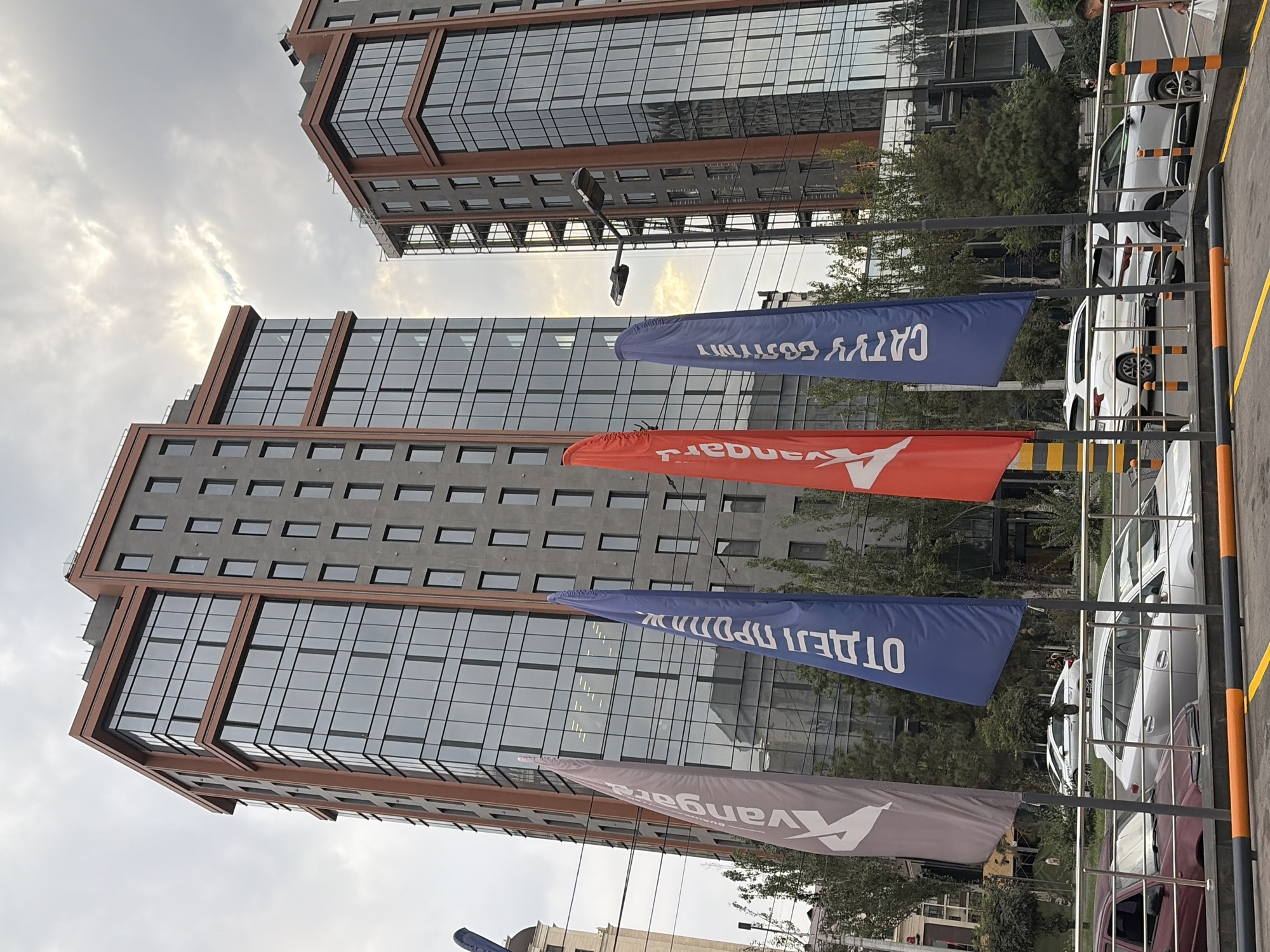Two modern, multi-story buildings with glass facades and reddish-brown trim stand prominently in the center and right of the frame. The building on the left is taller and wider than the one on the right. In the foreground, three colorful flags are planted: a light purple flag with white text, a blue flag with white text, and a red flag with white text. The flags are partially obscuring the lower floors of the buildings and a street with cars. Several cars are visible on the street, including a silver sedan in the middle ground and a dark red car in the foreground. Trees and greenery are interspersed between the buildings and along the street. The sky above is overcast with some patches of bright clouds. A street lamp is visible between the two buildings.
