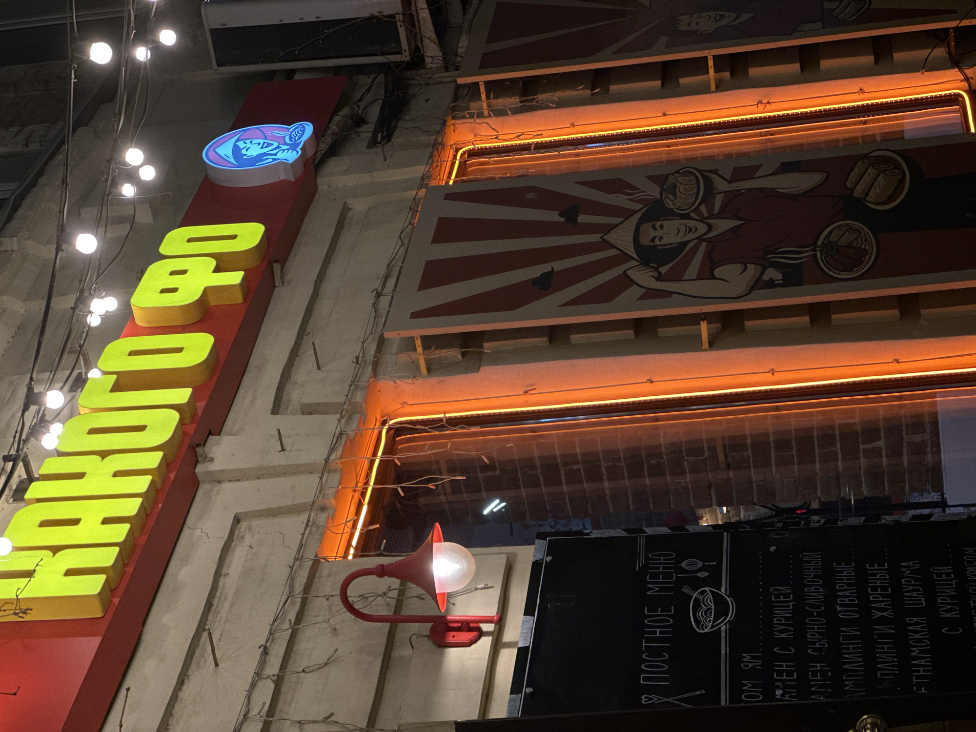 Here is a description of the image:
The image shows the exterior of a restaurant, likely in Rostov-on-Don, Russia, judging by the Cyrillic script on the signage. The sign reads "КАКОГО ФО," which possibly translates to "What Pho?" indicating a Vietnamese restaurant. The sign is vertical, with neon yellow letters on a red background and a blue logo at the top. Above the restaurant's main sign are decorative panels featuring cartoon-style illustrations of a person holding plates of food, illuminated by orange neon lights. A blackboard displays the menu in Cyrillic, listing various dishes, including ramen with chicken, dumplings, and Vietnamese shawarma with chicken. A red gooseneck lamp illuminates the menu board. The overall scene suggests a modern, urban setting, possibly a street with multiple food establishments.