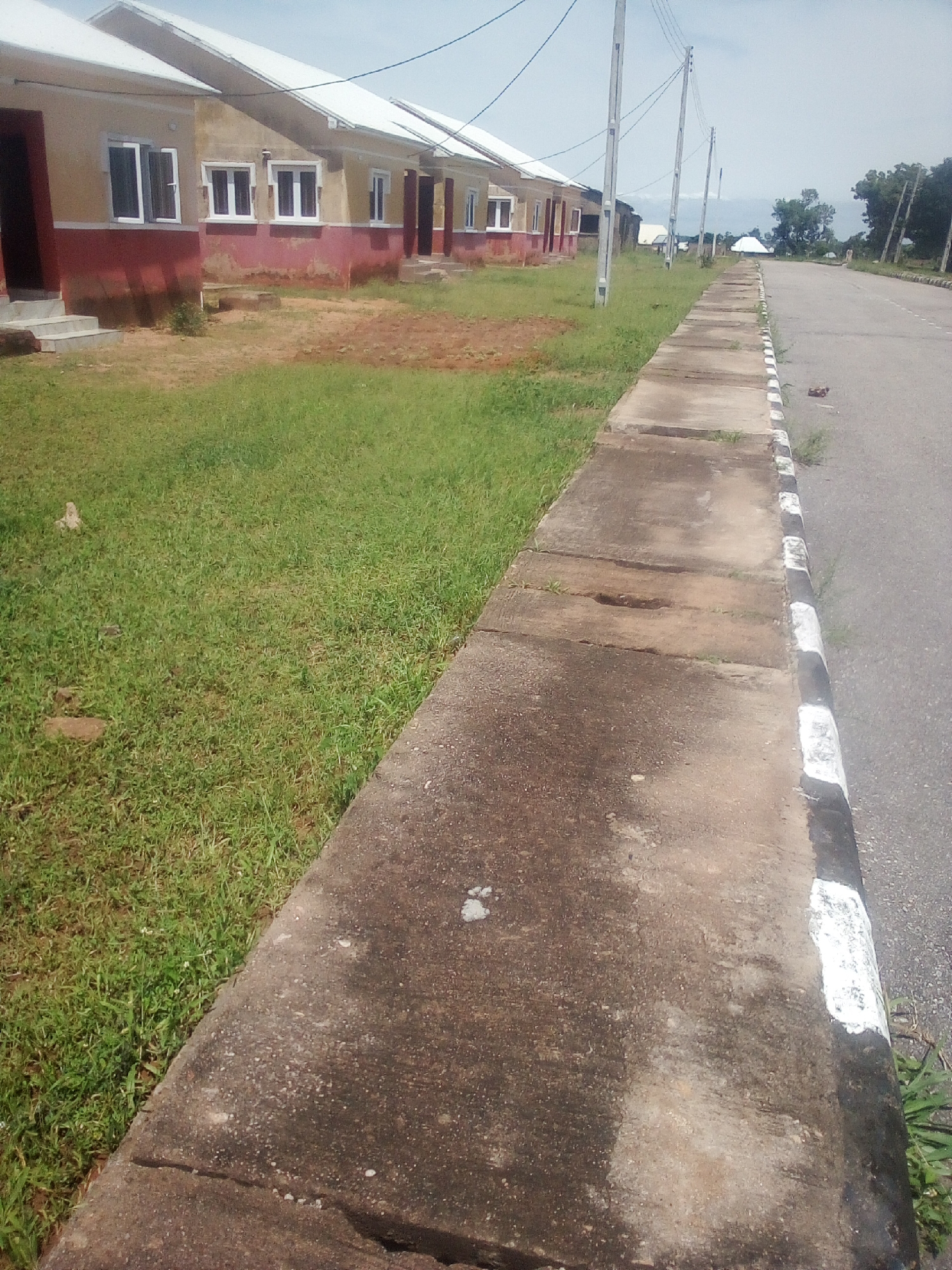 The image shows a row of identical, single-story houses with yellow and red facades, white trim, and light-colored roofs, located in Jauro Yinu, Nigeria. A concrete sidewalk runs along the right side of the houses, bordered by a paved road with black and white markings. Lush green grass covers the area between the houses and the sidewalk. Several utility poles with wires are visible in the background. The sky is overcast. The scene appears to be residential, with no people or vehicles present.