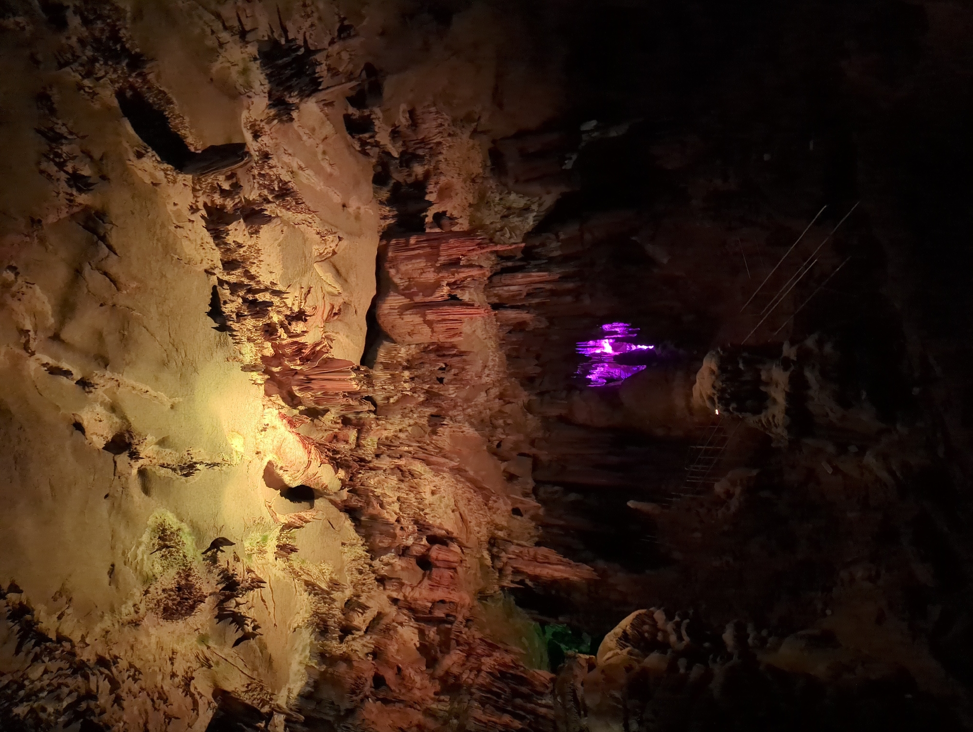 This is a photograph taken inside a cave. The cave is illuminated with artificial lights, casting warm tones of orange and yellow on the stalactites and stalagmites. Some areas are lit with a vibrant purple light, highlighting a particular section of the cave formations. Stairs with metal railings are visible, suggesting a path for visitors. The ceiling of the cave is covered in textured rock formations. The image is dark overall, with the lighting focusing on the geological features.