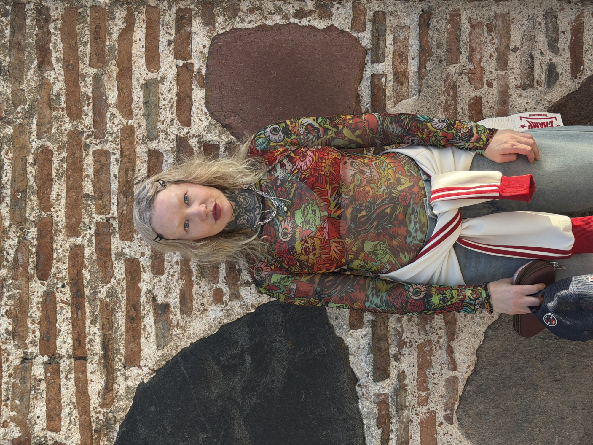 A woman lies horizontally on the ground in Hrodna, Belarus. Her head is to the left and feet to the right. She has blonde hair, blue eyes, and is wearing a sheer top with colorful skull and flower designs, a white jacket with red striped sleeves, and light blue jeans. She has dark tattoos around her neck and is wearing a silver necklace. A brown leather wallet and navy blue baseball cap sit near her right hand. Her left hand rests on her jeans. The ground appears to be an old brick and stone wall. A white box with the words "THE KNOCKOUT" and a star is partially visible next to her left hand.