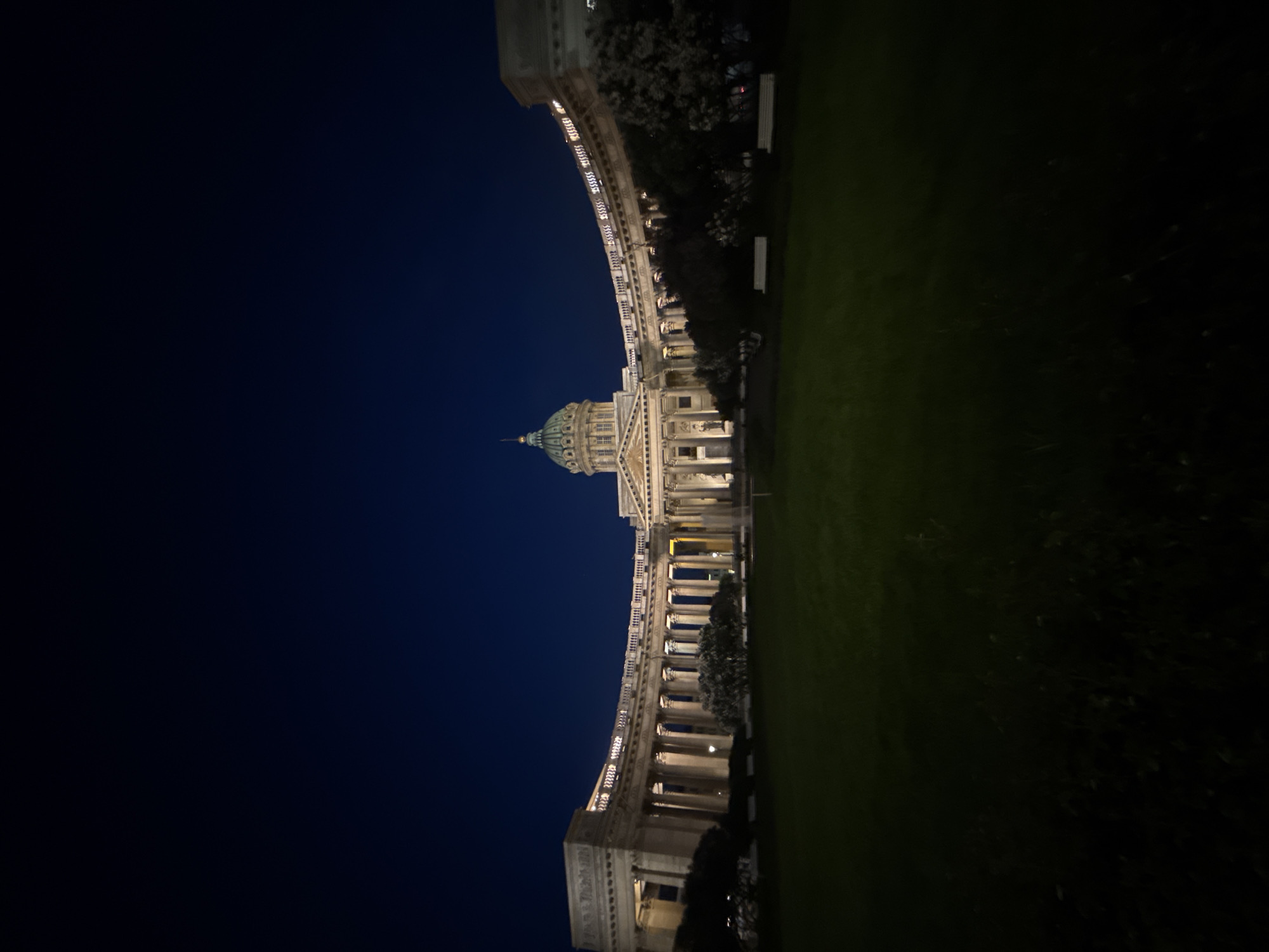The image displays a grand architectural landmark illuminated at night, strongly identifiable as the Kazan Cathedral in Saint Petersburg, Russia. The majestic building stands prominently, characterized by a large, green-domed rotunda topped with a lantern and cross, and a classical facade featuring a portico with multiple columns.
Extending from the main structure is a sweeping, semi-circular colonnade, also adorned with numerous columns, creating a dramatic curved architectural element. The entire building and its colonnade are brightly lit with artificial lights, highlighting the architectural details and the warm hues of the stone, contrasting sharply with the deep indigo night sky above.
In the foreground and to the right of the building, a dark green grassy lawn is visible, hinting at a public square or park area. Dark trees and foliage line the background and edges of the grassy expanse. The time of day is clearly night, evidenced by the dark sky and reliance on artificial lighting. No people or discernible activities are visible, lending a quiet and monumental atmosphere to the scene. There is no visible text in the image.
