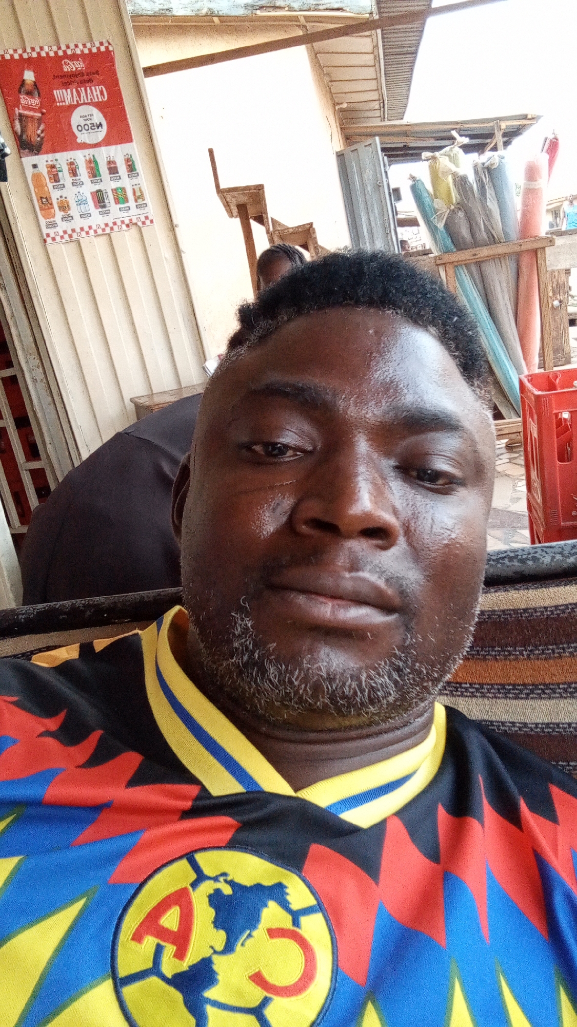 The image is a close-up selfie of an adult man with dark skin, a short, salt-and-pepper beard, and curly black hair. His face appears slightly sweaty. He is wearing a colorful sports jersey, predominantly black, with intricate red, yellow, and blue geometric patterns. A prominent logo featuring a yellow soccer ball with blue continental shapes and the red letters "A" and "C" is visible on the chest. The man looks directly at the camera with a neutral expression.
The setting is an informal outdoor or semi-outdoor commercial area, likely a market stall or small shop in Sabon Tasha, Nigeria. In the background, a person in a dark shirt is partially visible. To the left, an upside-down red and white advertisement poster is affixed to a light-colored corrugated wall. The poster displays a Coca-Cola bottle, other bottled and canned beverages, and prices like "N200" and "N300" (Nigerian Naira). Text such as "CHAYAH!!!" and other local script is also discernible. To the right, red plastic crates are stacked, and further back, rolls of various materials, possibly textiles, are stored upright in a wooden stand. The bright lighting suggests it is daytime.