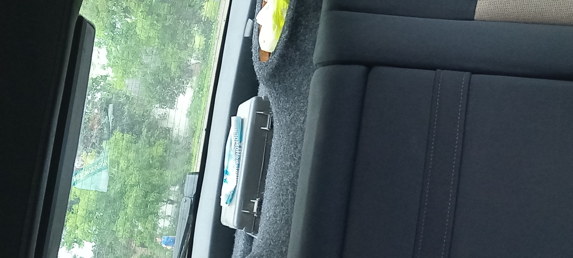 The image captures a view from inside a vehicle, likely a car or van, looking out the rear window. The setting appears to be Koviahy, Ukraine, with lush green trees and foliage visible outside, suggesting a daytime scene with moderate weather, possibly overcast or after rain due to the slightly muted lighting and visible streaks on the window.
Inside the vehicle, the focus is on the rear seating area. A portion of a gray fabric seat is prominent on the right side of the frame, featuring stitched detailing. To the left of the seat, tucked into a crevice, is a light gray or silver case, possibly a first-aid kit or tool box, with metal clasps. A blue and white item, possibly a packet of tissues or wipes with text in Cyrillic script, is placed on top of the case. A bright yellow item, possibly a piece of fabric or a bag, is partially visible near the top of the image, nestled against the car's interior lining.
On the rear window, a sticker or decal is affixed. It is predominantly light blue and green with some white elements. While the text is partially obscured and viewed through the glass, some Cyrillic characters are discernible, including what appears to be "ОРУЖІЖ" which translates to "weapons". The overall impression is one of practical preparedness within a vehicle, set against a natural, green backdrop.