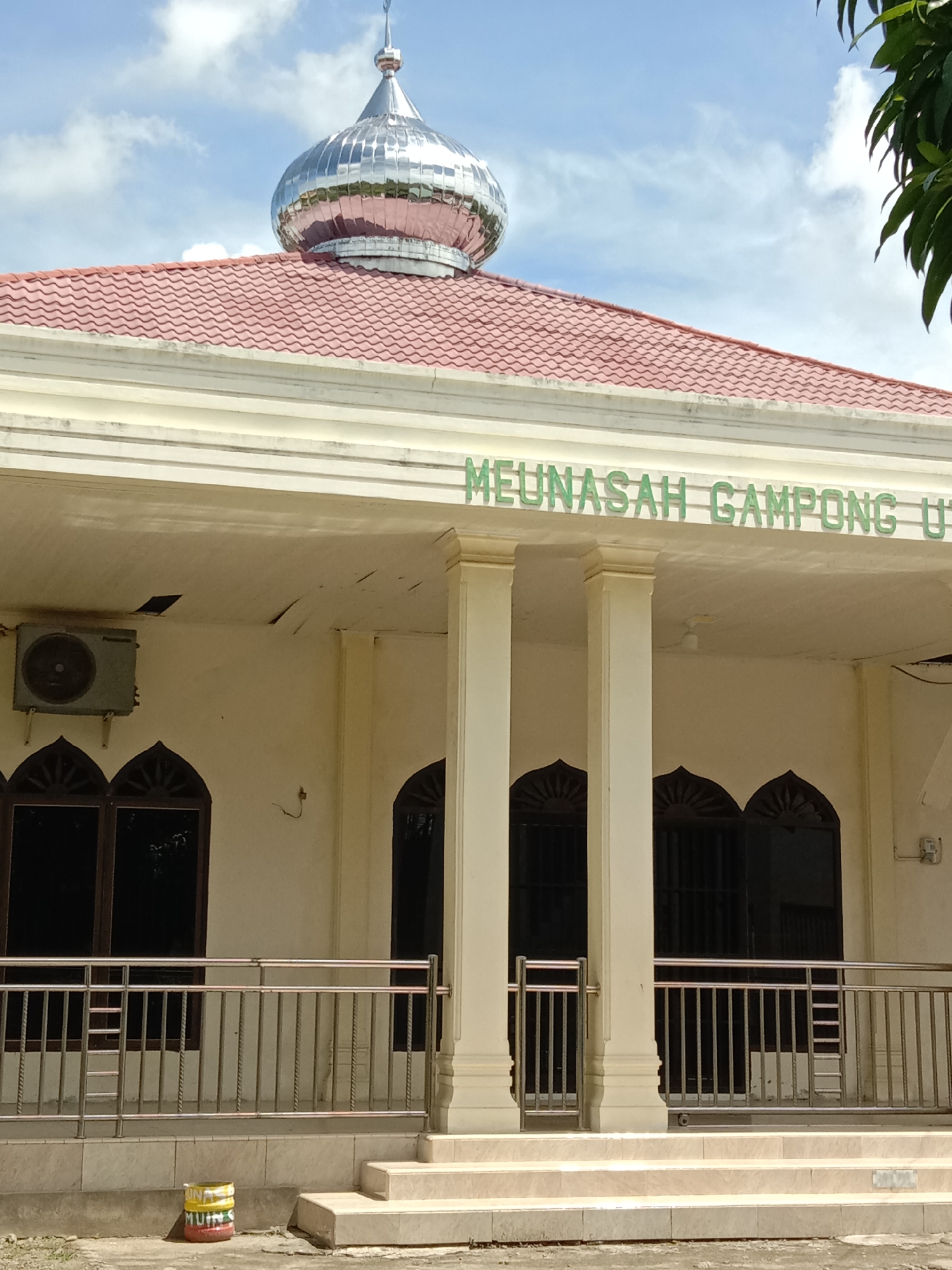The image depicts the Meunasah (a type of community hall or religious building) of Gampong Uteuen Bayi in Lhokseumawe, Indonesia. The building is a single-story structure with a light-colored exterior and a red tiled roof. A silver, onion-shaped dome sits atop the roof. The facade features a series of arched windows and doorways. A railing runs along the front of the building, and a set of stairs leads up to the entrance. An air conditioning unit is visible on the wall. A paint bucket sits on the ground near the stairs. The sky is visible in the background.