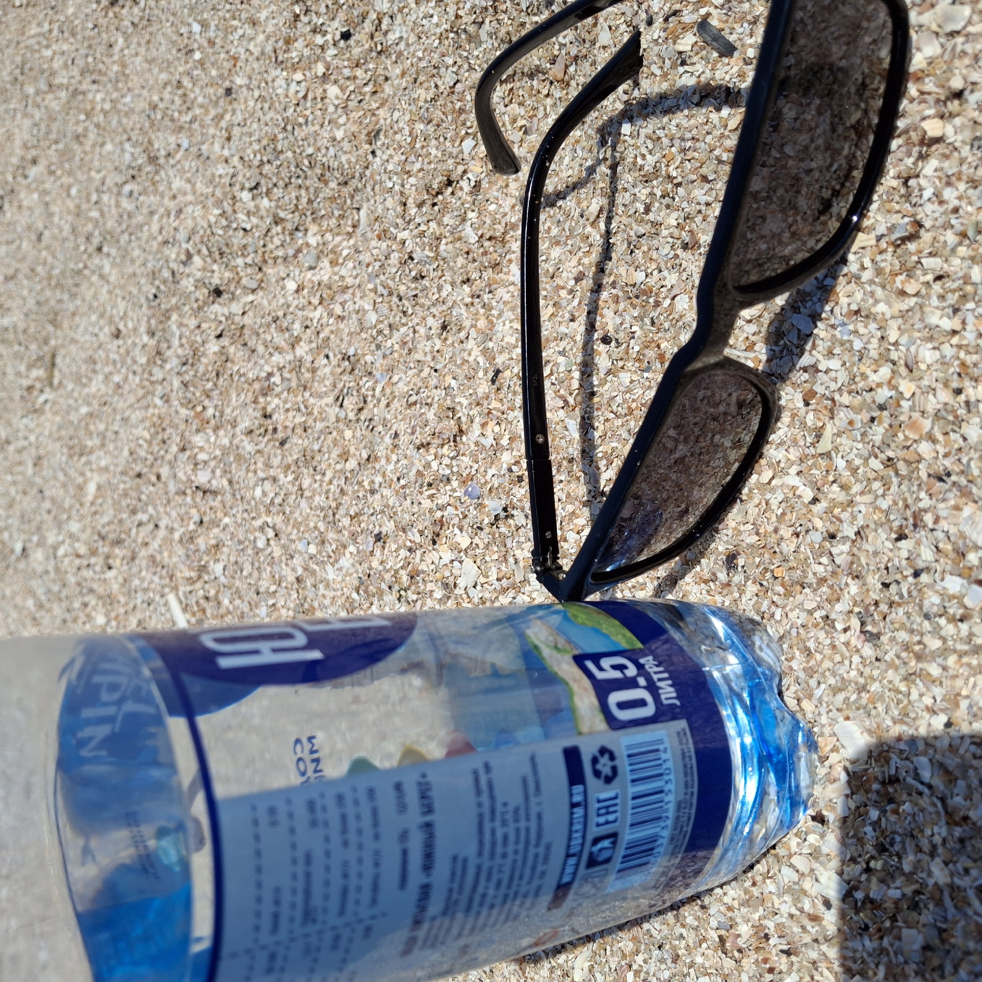 A discarded plastic water bottle and broken sunglasses rest on a beach composed of sand and shell fragments. The bottle is partially filled with liquid and labeled with Cyrillic text, along with the volume "0.5" and an "EAC" mark. The sunglasses are black-framed with tinted lenses; one arm is detached from the frame. The location is Unknown, Unknown.