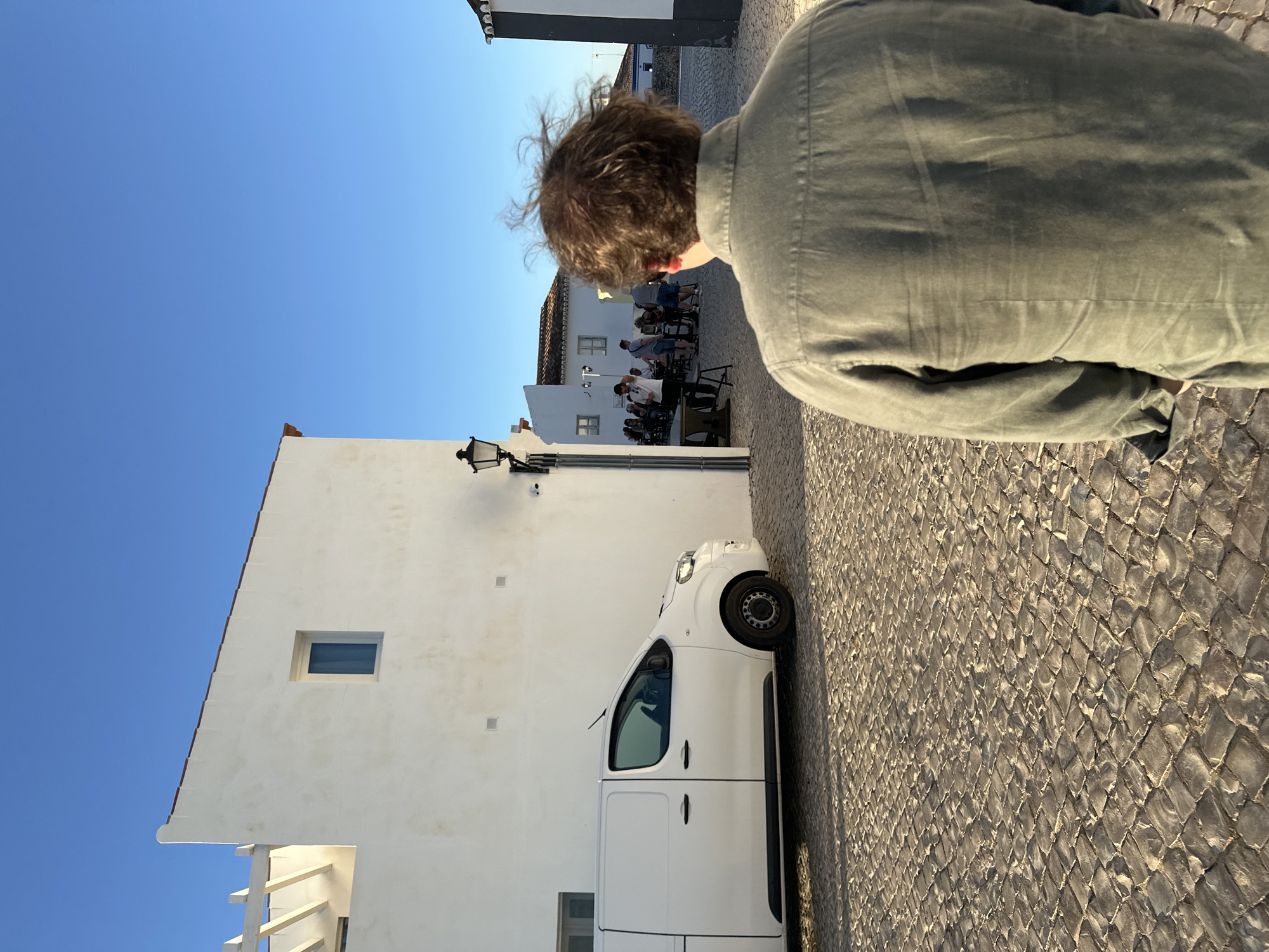 A man stands on a stone-paved street in Vila Real de Santo António, Portugal, his back to the camera. To the left, a white building with a window and a parked white van is visible. A group of people are seated at tables in the background. The sky is blue.