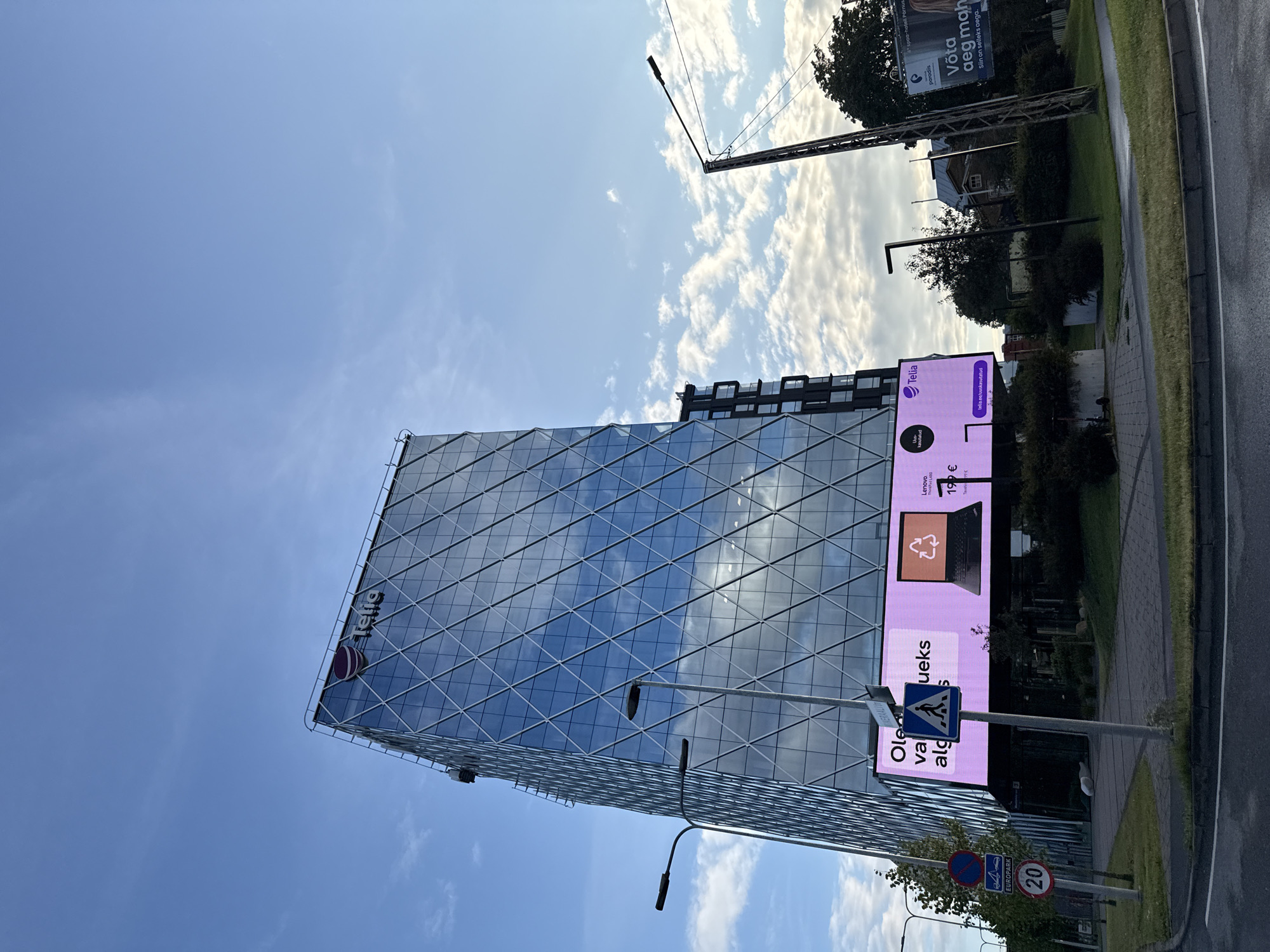 The image shows a modern glass-fronted building in Tallinn, Estonia, with the Telia logo visible at the top. A large digital billboard is displayed on the side of the building, advertising a Lenovo laptop with a recycling symbol and a price of 199 €. The billboard also features the Telia logo and a website address. In the foreground, there are streetlights, traffic signs including a speed limit sign indicating 20 km/h, and a pedestrian crossing sign. The sky is blue with scattered clouds, and there is green grass and trees around the building, suggesting an urban environment. The overall scene depicts a commercial area in the city.