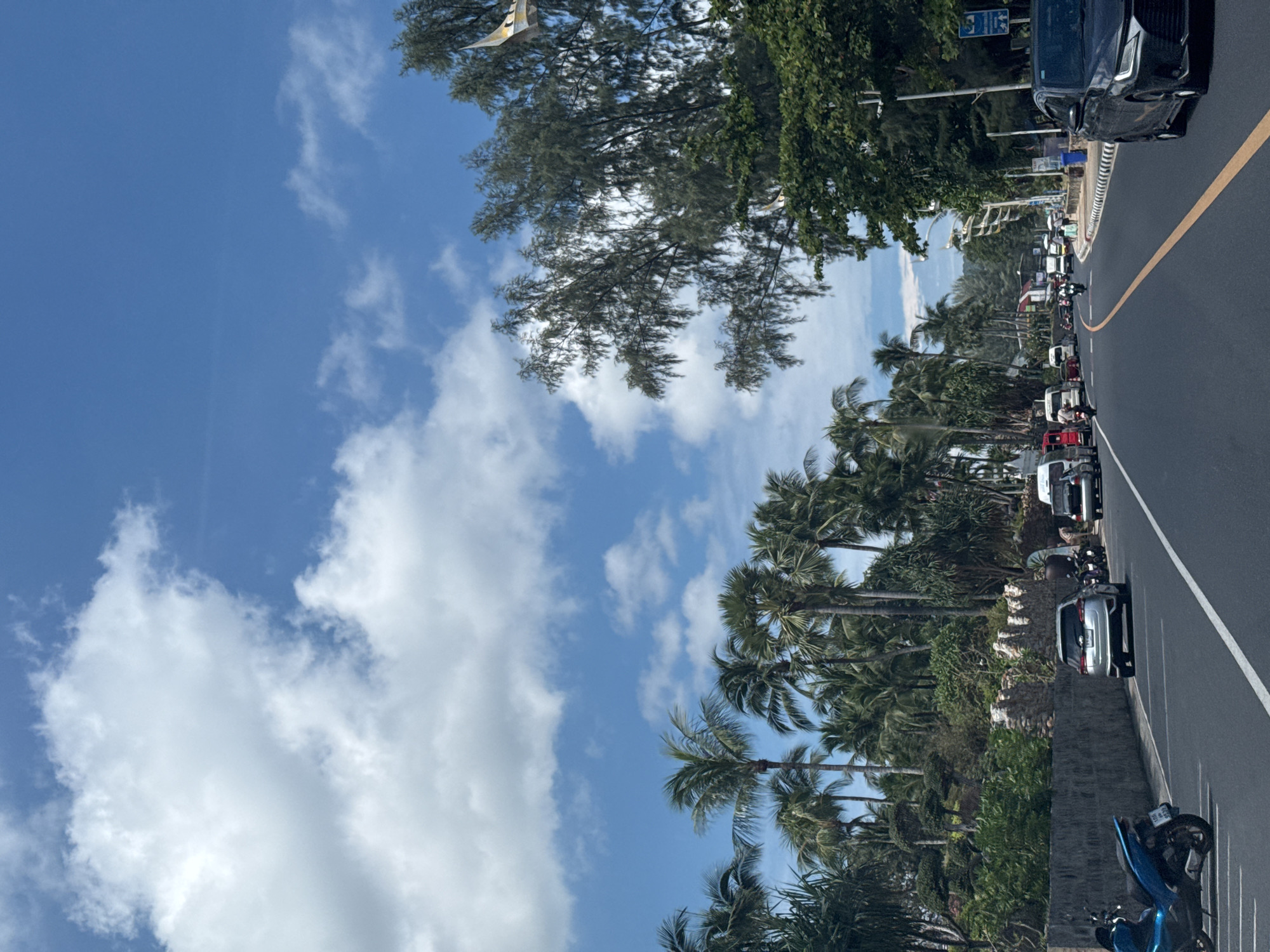 The image depicts a street scene on a sunny day with a clear blue sky and scattered white clouds. Lush palm trees and other greenery line the left side of the road, interspersed with a stone wall. A variety of vehicles, including cars, motorcycles, and vans, are present on the asphalt road. Some people are visible walking on the sidewalk on the right side of the road. The overall impression is of a tropical urban environment.