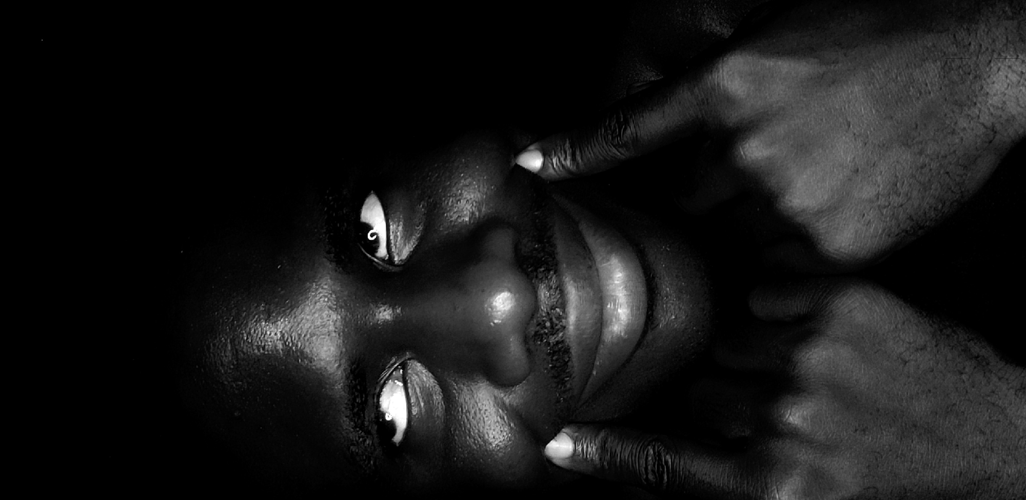 The image is a monochrome close-up portrait of a person of African descent. The person's face is the main subject, positioned at a tilted angle. Two hands are partially visible, each with a finger pointing towards the person's cheeks. The setting is dark. The image provides no evidence that the scene is located in the city of Narayi, Nigeria.