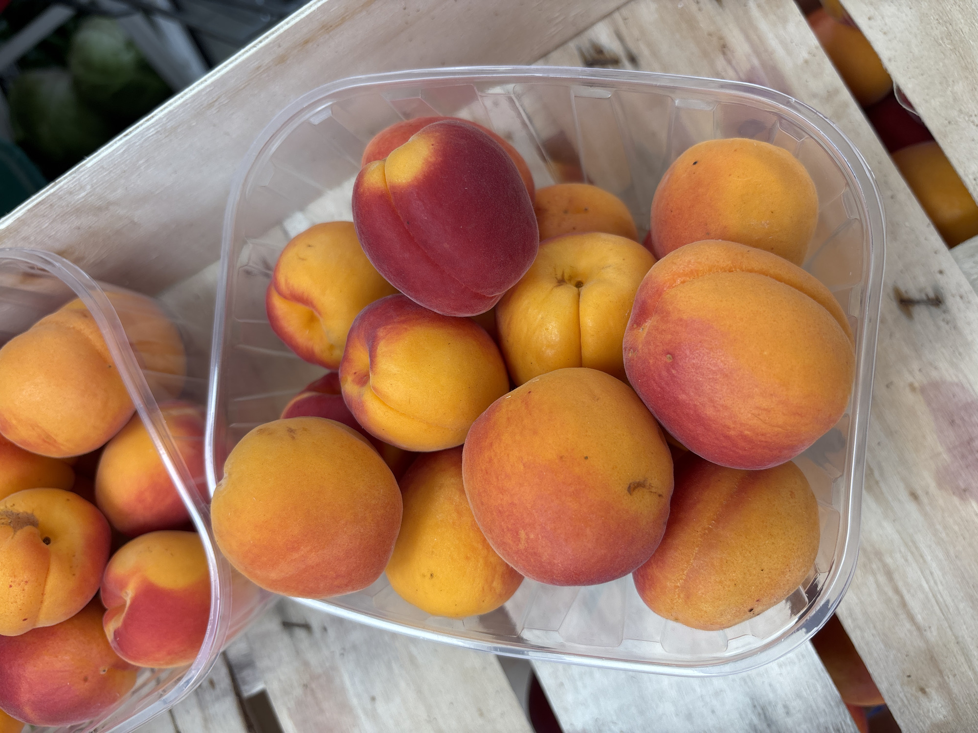 This is a close-up, eye-level, full-frame shot of two clear plastic containers filled with ripe apricots, placed on a light-colored wooden surface. The apricots are predominantly orange with some exhibiting a blush of red. Some fruits have visible small dark spots. The containers are positioned to overlap each other in the frame. The background is out of focus, showing hints of green and what appears to be a wooden crate. The scene suggests a market or a stall, possibly in the city of Boreti, Montenegro, where fresh produce is being sold. There are no people visible in the image.