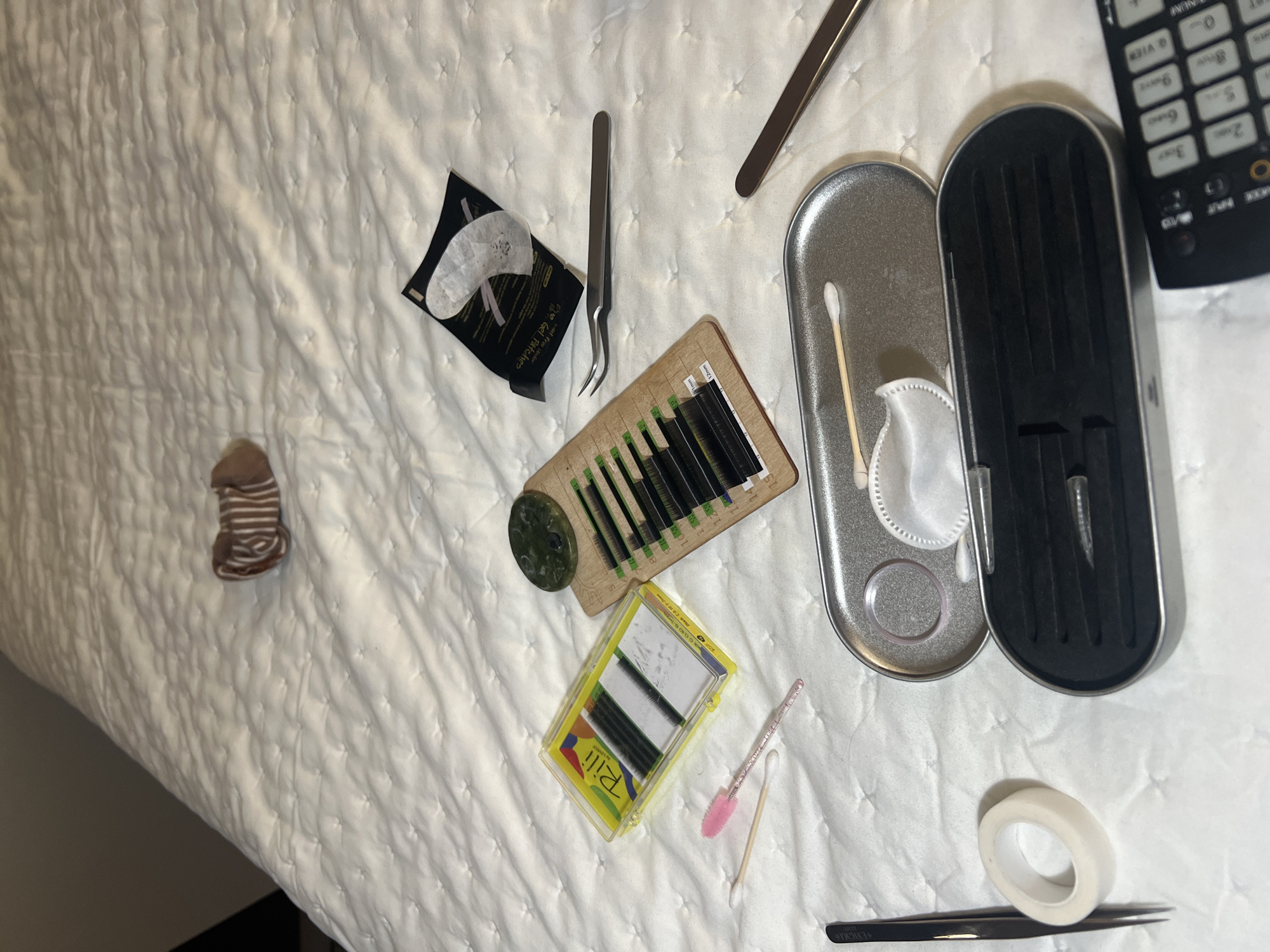 The image shows an overhead shot of various beauty-related items arranged on a white, quilted bedspread. These items include: eyelash extensions in multiple cases, tweezers, cotton swabs, eye gel patches, a small roll of tape, and a small, striped object appearing to be a sock or a similarly patterned item. A remote control is visible on the upper right. The scene seems to depict items related to eyelash extension application. The location is not discernible from the image.