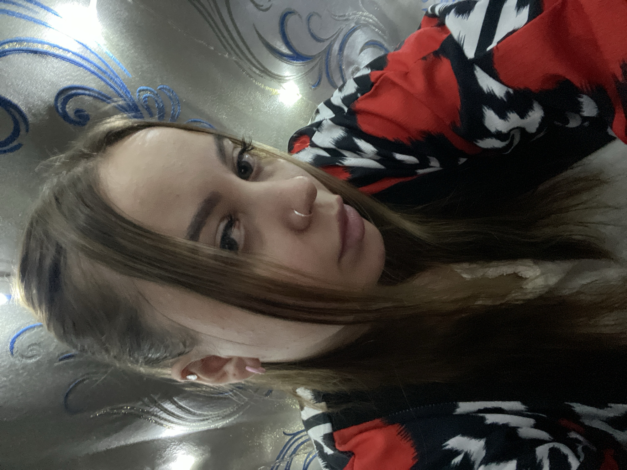 A young woman with light brown hair and fair skin is lying down, facing the camera. She has a nose ring. Her clothing is a red, black, and white patterned garment. The background appears to be a patterned surface, possibly fabric or wallpaper, with blue and silver elements. No specific location in Novopokrovskaya, Russia can be determined from the image.