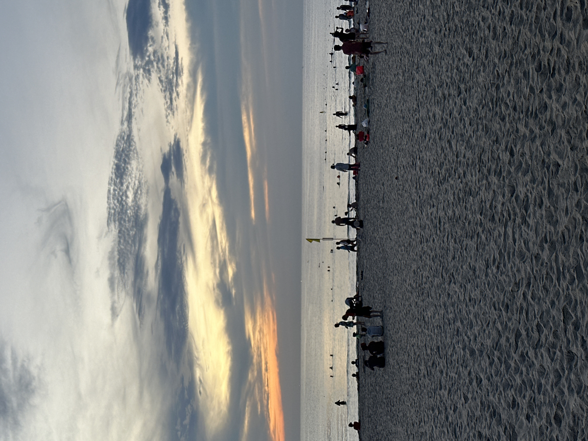 This image depicts a beach scene at sunset in Da Nang, Vietnam. The sky is a gradient of colors, with shades of orange, yellow, and gray from the setting sun blending into the clouds. The ocean is calm and stretches to the horizon, where a faint line of distant lights can be seen. The foreground is a sandy beach populated by numerous people engaged in various activities. Some individuals are wading in the shallow water, while others are standing or sitting on the sand. A group of people are gathered near the waterline, and further up the beach, more individuals are visible, some appearing to be walking or relaxing. There are no discernible structures or buildings in the immediate vicinity, emphasizing the natural coastal environment. The overall impression is of a serene evening at a popular beach destination.