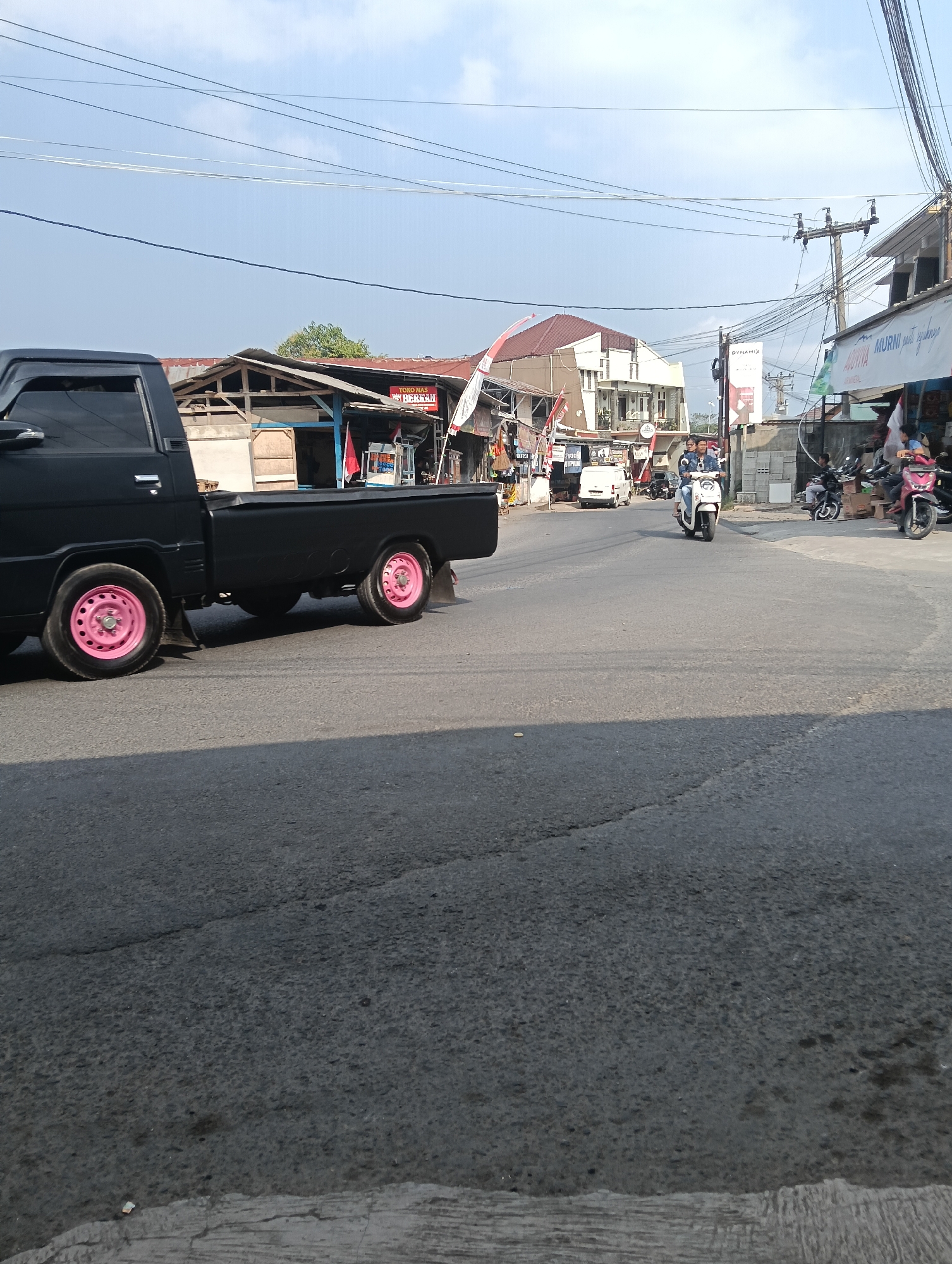 The image depicts a street scene in Puspahiang, Indonesia, under a partly cloudy sky. A black pickup truck with pink rims is parked on the left side of the road, its rear facing the viewer. The truck is positioned on an asphalt road. In the background, various buildings line the street, including shops with awnings and signage. One shop displays the text "TOKO MAS BEKALI". Several motorcycles are visible on the road and parked near the shops. Two people are riding on a white scooter in the middle of the road. Further down the street, a white van is also visible. Utility poles with numerous electrical wires are present on the right side of the image. The overall environment suggests a commercial area within the town.
