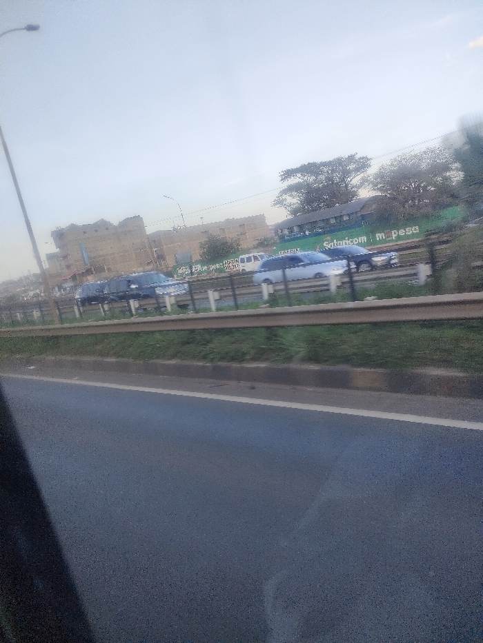 The image depicts a daytime outdoor scene on a road in Theta ward, Kenya, under an overcast sky. In the immediate foreground, the dark interior frame of a vehicle partially obscures the view of an asphalt road with a white edge line. A grassy median strip with a brown curb and a metal guardrail separates this foreground element from a multi-lane asphalt road to the right. On this road, several vehicles are visible: a dark SUV or sedan, a light-colored sedan, another dark vehicle, and a white van in the distance, all appearing to be in traffic. In the midground, a large green billboard with white lettering clearly displays "Safaricom m-pesa," along with other text including "RAZIM" and "PEMBA DEBA." To the left of the billboard, multiple multi-story buildings, some with incomplete exteriors and one with a crenellated roofline, are present. To the right of the billboard, several trees with dense foliage are visible. A tall street light pole with a single luminaire extends from the lower left towards the top center. Another smaller street light is visible further back among the buildings.