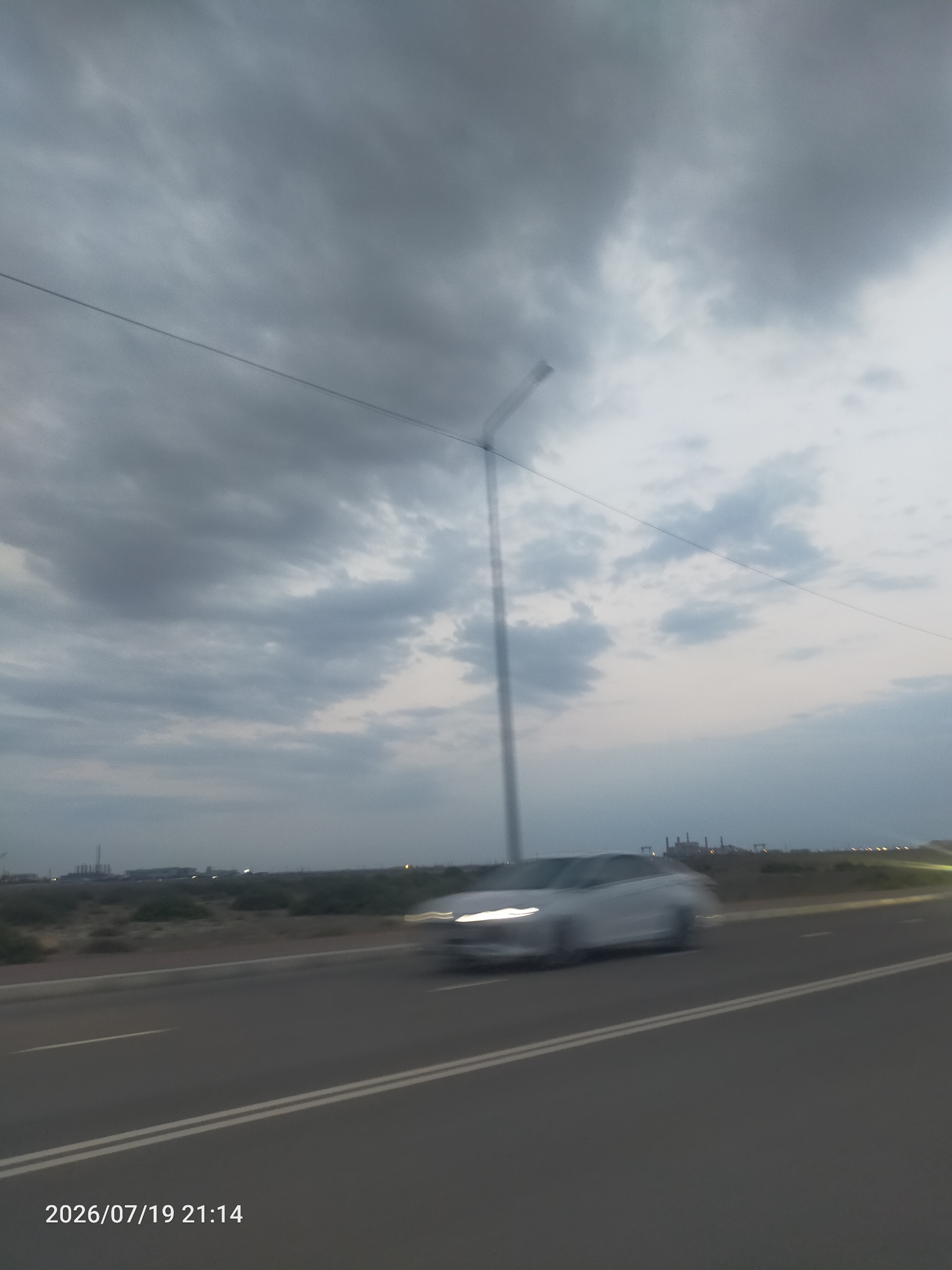 The image depicts a road in an industrial area at dusk. A car is captured in motion on the left side of the road, appearing slightly blurred due to its speed. The sky is overcast with dark clouds. In the background, industrial structures are visible on the horizon, along with a series of lights. The road is paved and has white lane markings. The timestamp "2026/07/19 21:14" is present in the bottom left corner of the image.