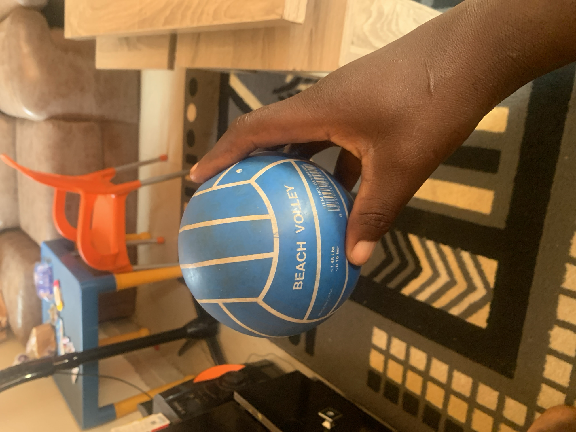 The image shows a hand holding a blue and white volleyball. The volleyball has the words "BEACH VOLLEY" printed on it, along with some smaller text and a barcode. The background is blurred, but it appears to be an indoor setting with furniture, including a couch and a table. The image does not provide any information about the location in the city of Gura Top, Nigeria.