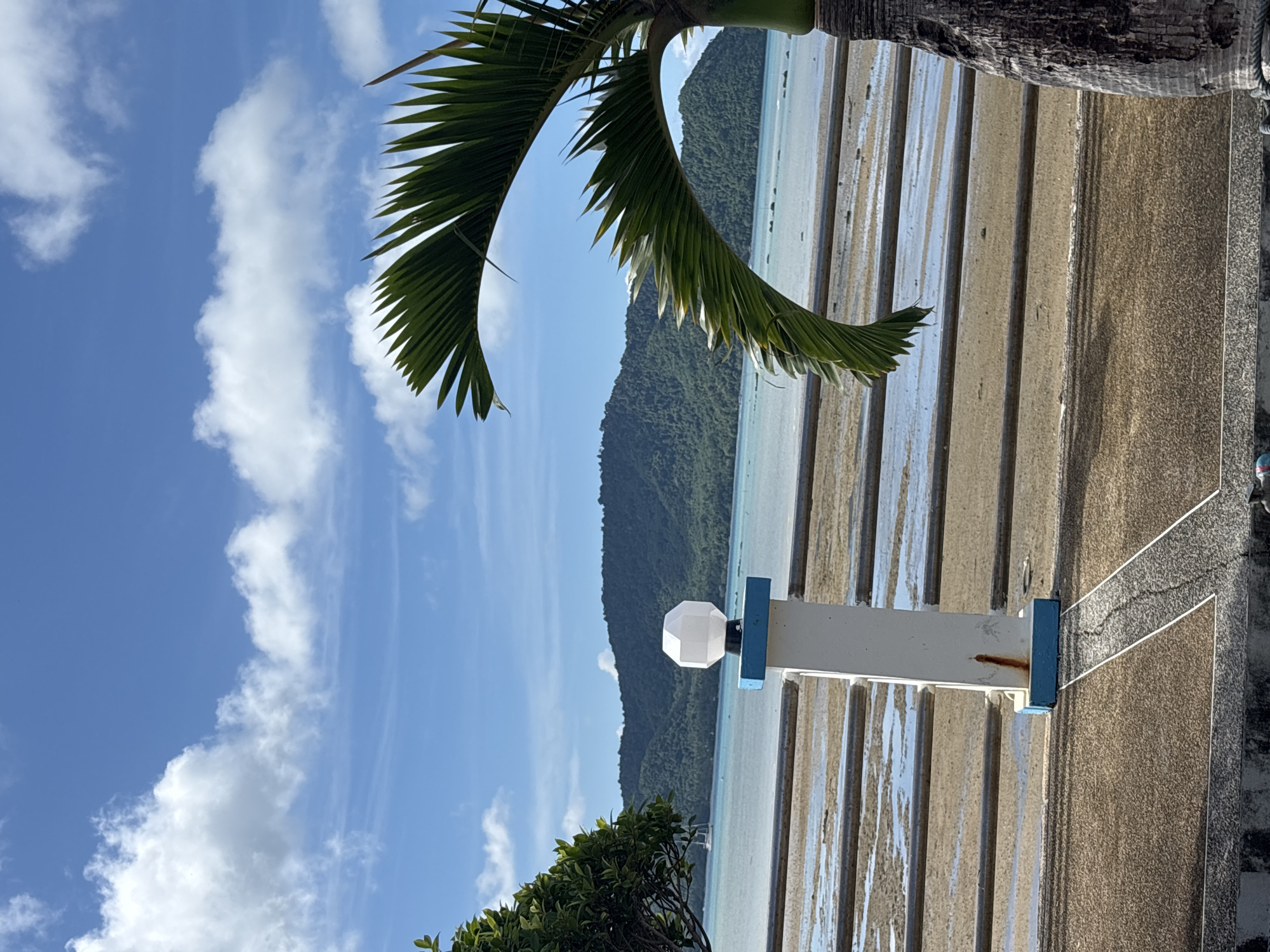 The image depicts a scenic view of Rawai, Thailand, featuring a coastal landscape under a bright, partly cloudy blue sky. In the foreground, a white post with a blue base and a geometric white light fixture stands beside a palm tree. A metal railing separates the paved area from a sandy, wet beach with shallow water. In the mid-ground, the calm sea stretches towards lush, green, tree-covered hills in the background. A solitary sailboat is visible on the water. The overall scene suggests a peaceful, tropical environment.
