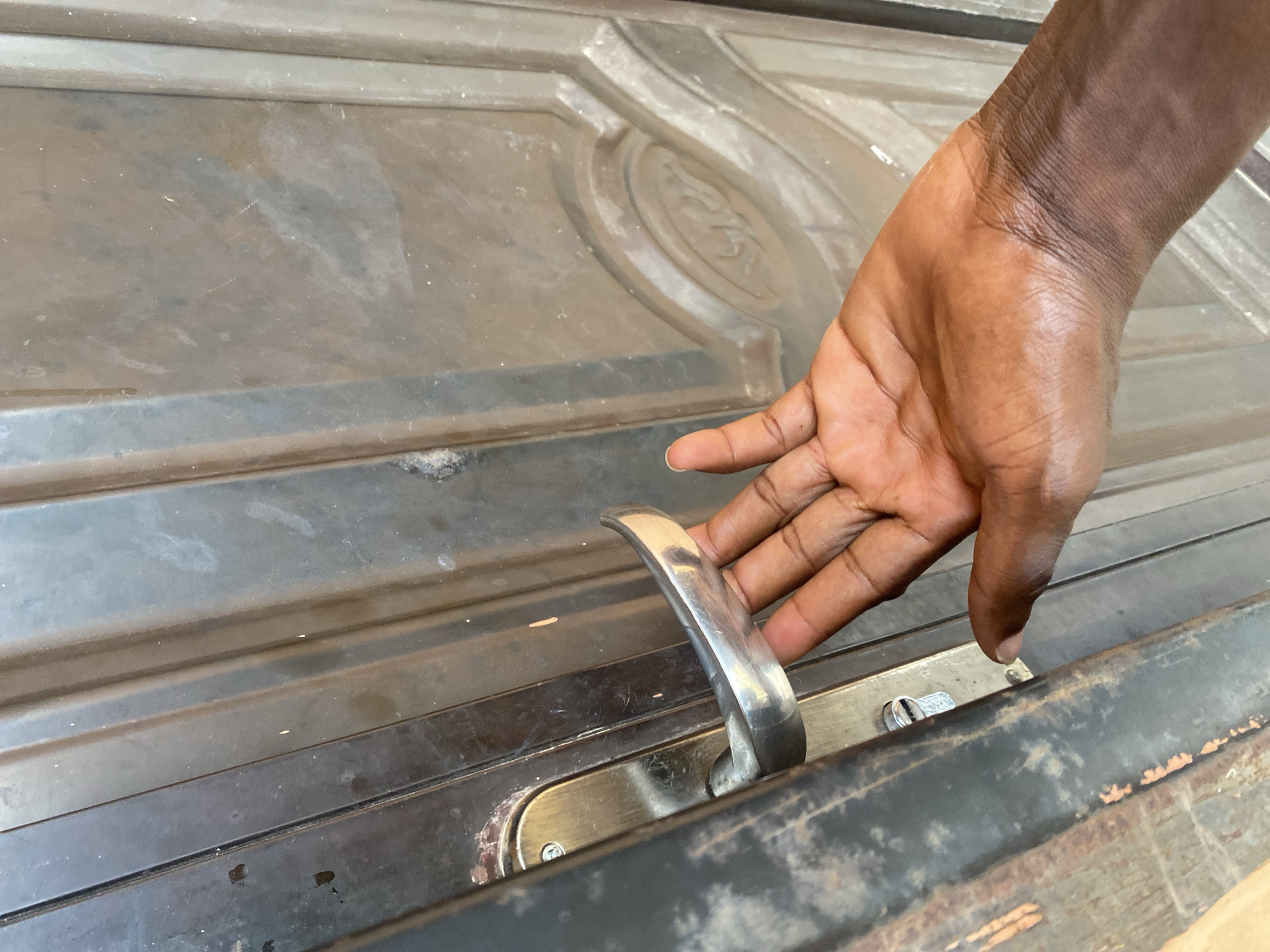 A close-up shot focuses on a person's hand reaching for the handle of a weathered, dark-colored metal door. The hand, with its dark skin and visible creases, is positioned to grasp the curved, silver door handle. The door itself appears to be made of metal and has a raised, decorative panel with a circular design in the upper right portion of the frame. The surface of the door is dusty and shows signs of age and use, with subtle scratches and smudges. Below the handle, a lock mechanism is visible, with a screw head and a keyhole. The scene appears to be outdoors, given the texture and wear of the door, and the lighting suggests it could be daytime. The image captures a simple, everyday action of opening or closing a door. The setting is identified as Bulamare, Nigeria. There is no visible text in the image.