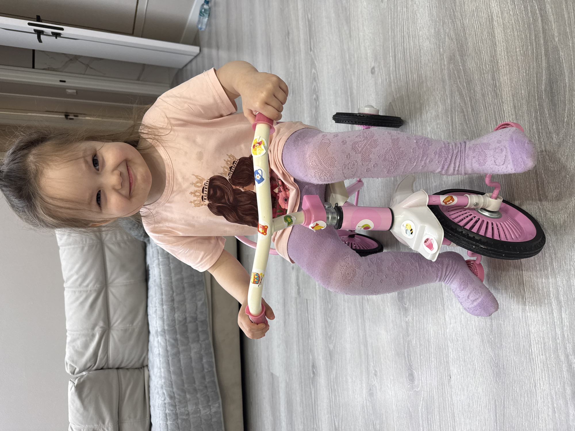 A young girl is captured indoors, straddling a pink balance bike and smiling directly at the camera. She appears to be a toddler, with light brown hair and a visibly cheerful expression. She is dressed in a light pink t-shirt featuring a graphic of two girls with long dark hair and crowns, paired with light purple patterned tights. Her hands are holding the yellow handlebars of the balance bike, which are adorned with various colorful stickers depicting objects like a drum, a bee, and a teacup, as well as cartoon characters.
The balance bike has a pink and white frame and pink wheels, consistent with a child's toy. The setting is an interior space, likely a home, with light gray wood-look laminate flooring. To the left, a portion of a light gray quilted sofa or couch is visible. In the background, there is a white cabinet or furniture piece with black handles and a light-colored countertop, possibly marble-patterned. A plastic water bottle rests on this surface. The lighting suggests it is daytime. The image conveys a happy and playful moment, focusing on the girl's interaction with her bike. There is no discernible text in the image.