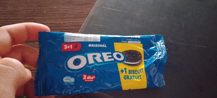 A hand holds a blue Oreo cookie packet. The packet is labeled "ORIGINAL" and includes a picture of an Oreo cookie. It is also labeled "3+1" and "+1 BISCUIT GRATUIT" with a yellow background. Nutritional information is visible at the top. The packet also indicates "2 dhs+." The packet is positioned on a wooden surface and a dark surface.