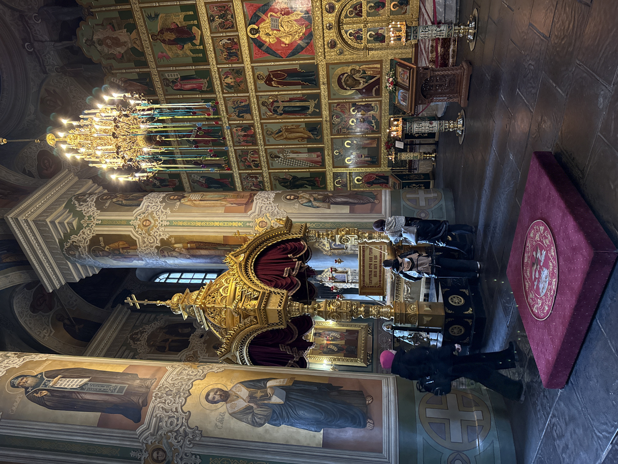 The image captures the interior of a Russian Orthodox church, possibly the Kazan Cathedral in Kazan, Russia, given the architectural style and iconography. The scene is ornate, with an emphasis on gold and religious artwork.
A large, multi-tiered golden chandelier dominates the upper left of the frame. The ceiling is covered with painted icons, each framed in gold. Walls are adorned with religious figures in traditional Orthodox style.
In the center, there's a golden structure, possibly a reliquary or shrine, draped with red fabric. A group of people, some wearing dark clothing and one in a pink hat, are present. They appear to be tourists or pilgrims, possibly viewing or paying respects at the shrine. Some appear to be praying.
The floor is made of dark stone tiles. To the right, there is a burgundy cushion with a gold emblem on it. Additional religious items, such as candlesticks and smaller icons, are placed around the shrine. The overall atmosphere is solemn and reverential.