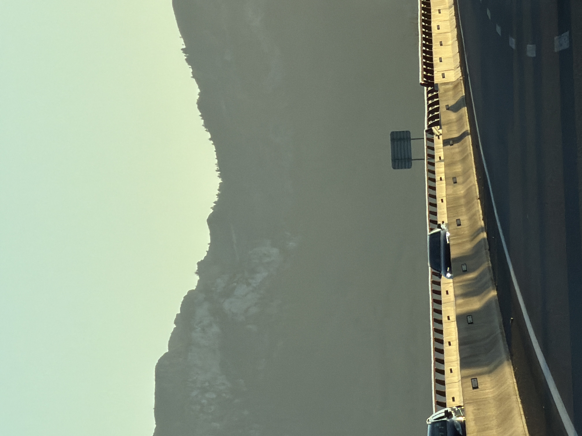 The image shows a highway in Mont-roig del Camp, Spain, with mountains in the background. The sky is a pale blue and the mountains are silhouetted against it. There are cars driving on the highway, and a concrete barrier separates the lanes. The sun is low in the sky, casting long shadows.