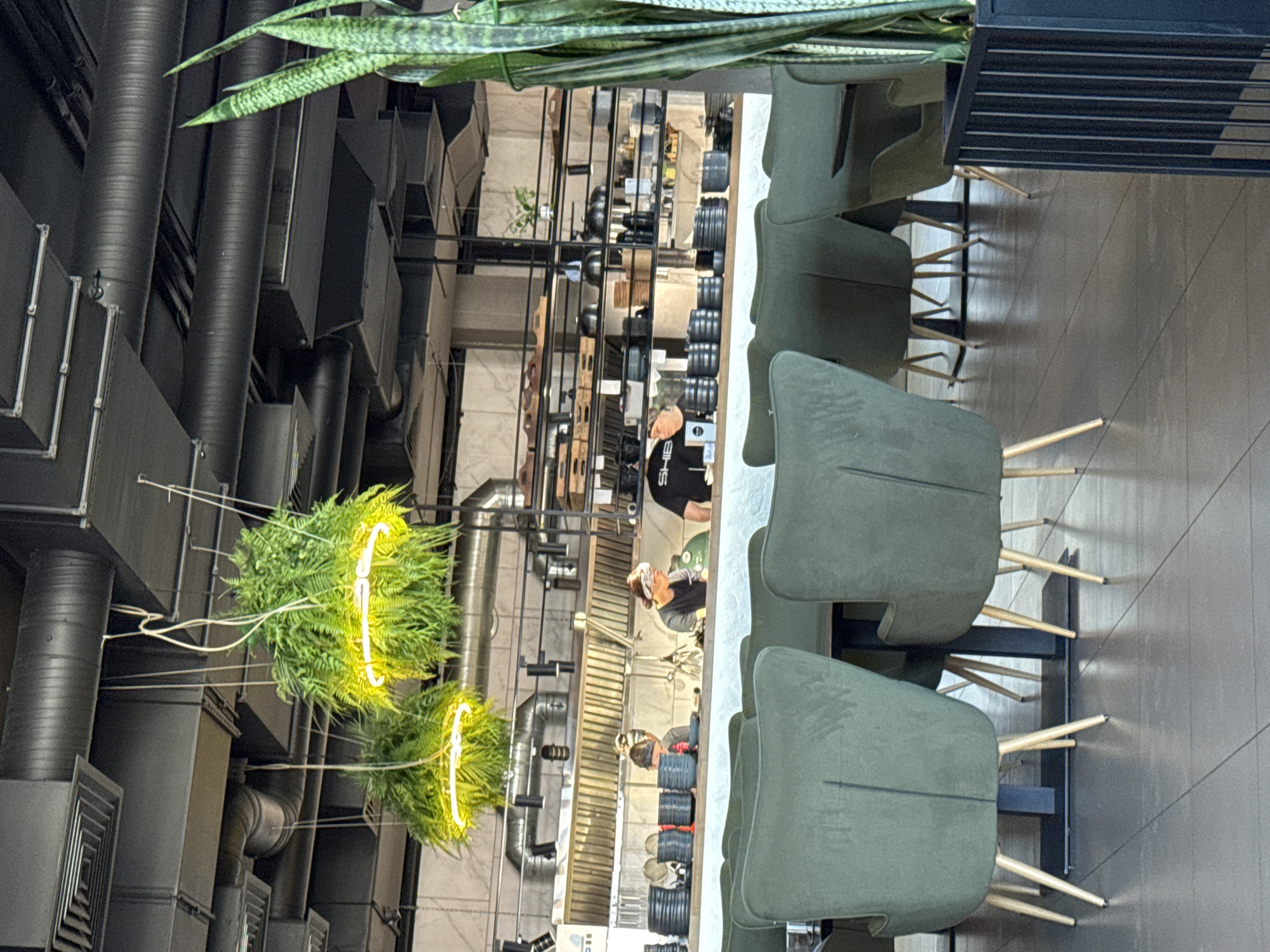 The image depicts the interior of a modern restaurant with an open kitchen. In the foreground, several plush green armchairs with light wooden legs surround dark-topped tables. To the right, a tall snake plant in a black planter adds a touch of greenery. The background reveals the bustling kitchen area, with stainless steel surfaces and shelves stocked with dishes and cooking utensils. Two individuals, presumably kitchen staff, are visible working behind a counter. One is wearing a black t-shirt with the word "SHIB" printed on it and a bandana. The other person, to their left, is also wearing a bandana and appears to be engaged in food preparation. Hanging from the ceiling are two large, spherical plant installations with integrated yellow neon lighting. The restaurant's infrastructure, including exposed ventilation ducts and industrial-style lighting, is visible on the ceiling. The overall ambiance suggests a contemporary dining establishment.