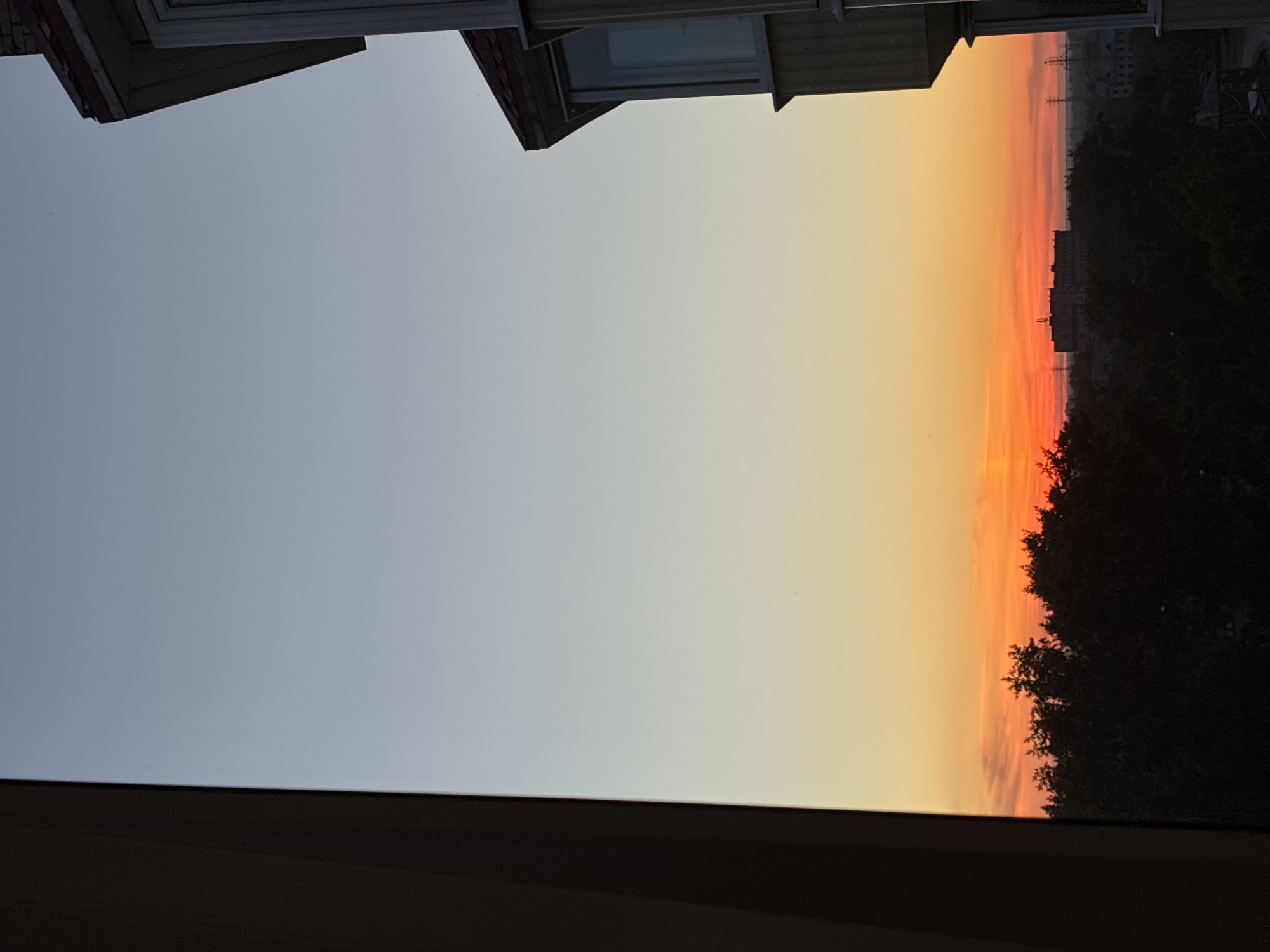 The image captures a sunset over an urban landscape. The sky transitions from a pale blue at the top to a vibrant orange and red glow near the horizon. Silhouetted against the colorful sky are the dark shapes of trees, forming a dense canopy across the middle ground. Beyond the trees, several buildings are visible, including a large, multi-story structure with antennas on its roof and smaller, industrial-looking buildings with power lines. The right side of the frame features a portion of a building with a visible window and a red-tiled roof, suggesting a residential or commercial structure. The left side of the frame is dominated by a dark, vertical element, possibly a window frame or part of a building's facade, that partially obstructs the view of the sunset. The overall scene suggests a view from a window looking out over a city at dusk.