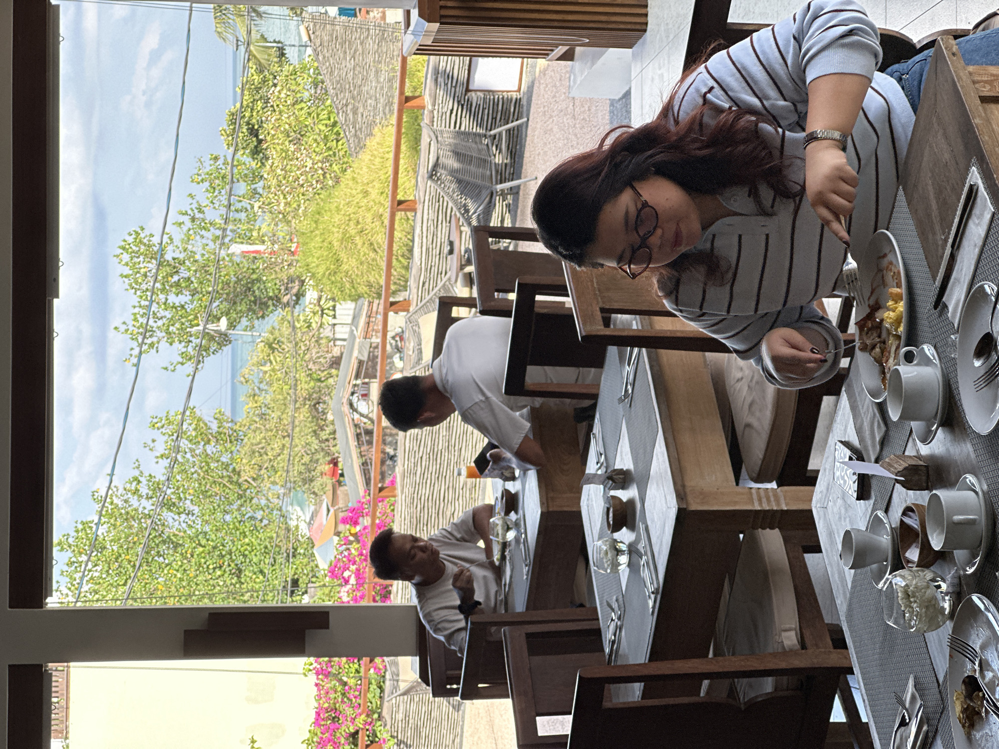 In Kuta, Indonesia, an outdoor seating area of a restaurant is depicted, overlooking a lush, tropical environment with the ocean visible in the distance. Three individuals are seated at tables, engaged in dining. In the foreground, a woman with dark hair and glasses is wearing a striped long-sleeved shirt and blue jeans. She is actively eating, using a fork and knife to cut food on her plate. To her left, a table is set for dining with plates, cups, and cutlery. In the middle ground, two men are seated at a table. One man, wearing a light-colored t-shirt, is looking down at his food and appears to be eating. The other man, wearing a white shirt, is partially visible from behind and is looking down. Behind the tables, a railing separates the dining area from an outdoor patio with chairs and tables. The background reveals vibrant pink bougainvillea flowers, green trees, and a glimpse of the ocean under a cloudy sky. The architecture includes wooden structures and tiled roofs, suggesting a resort or hotel setting.